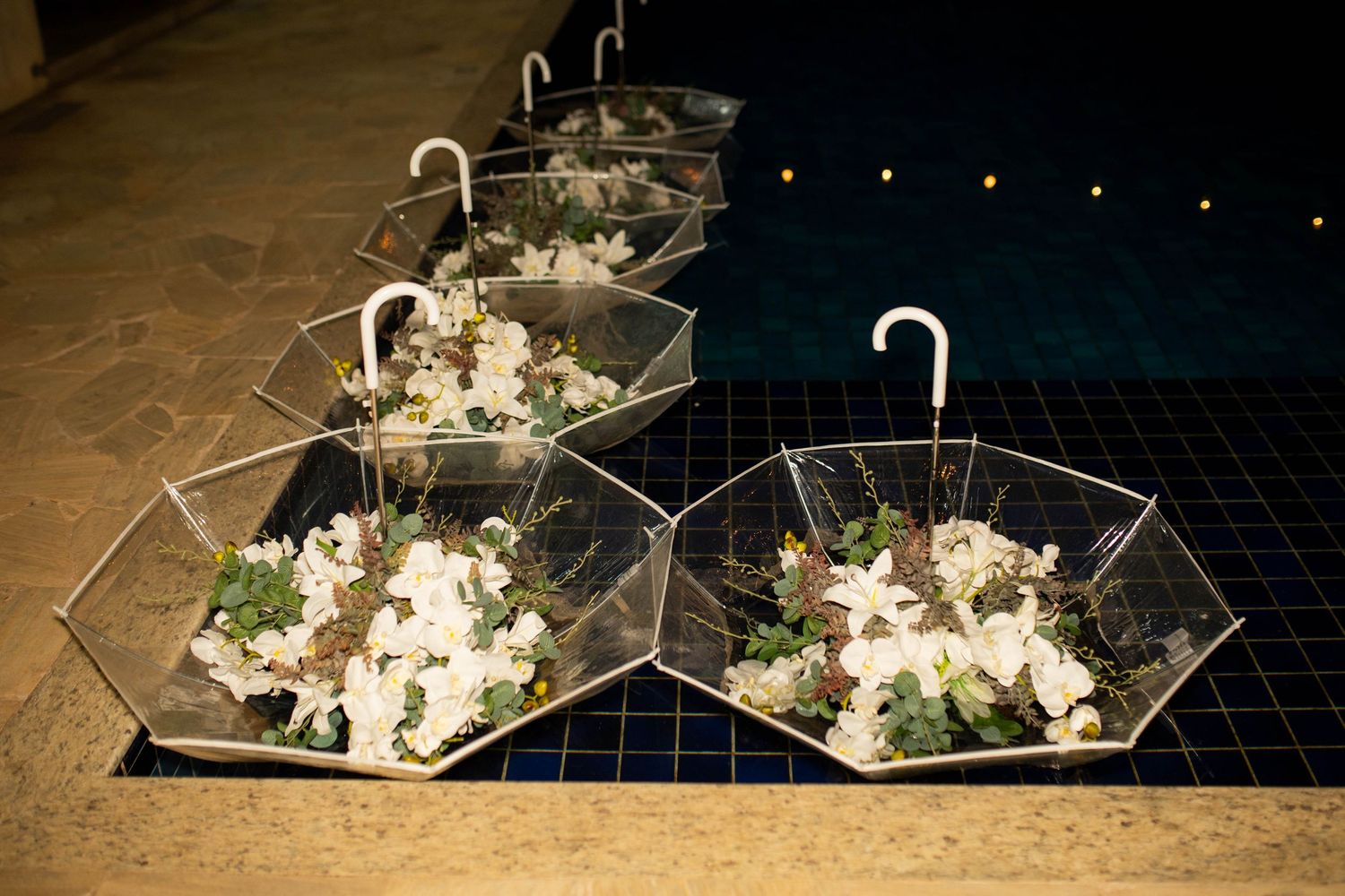 Clear umbrella centerpieces filled with white flowers and greenery displayed near a pool at an elegant event.