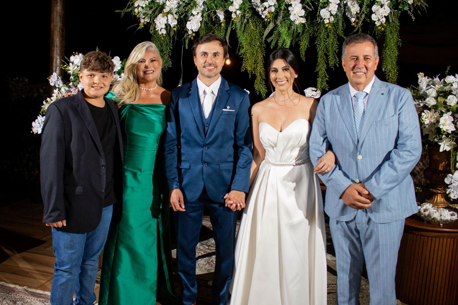 A joyful wedding party of five poses together, with the bride in white and groom in navy, flanked by family members.