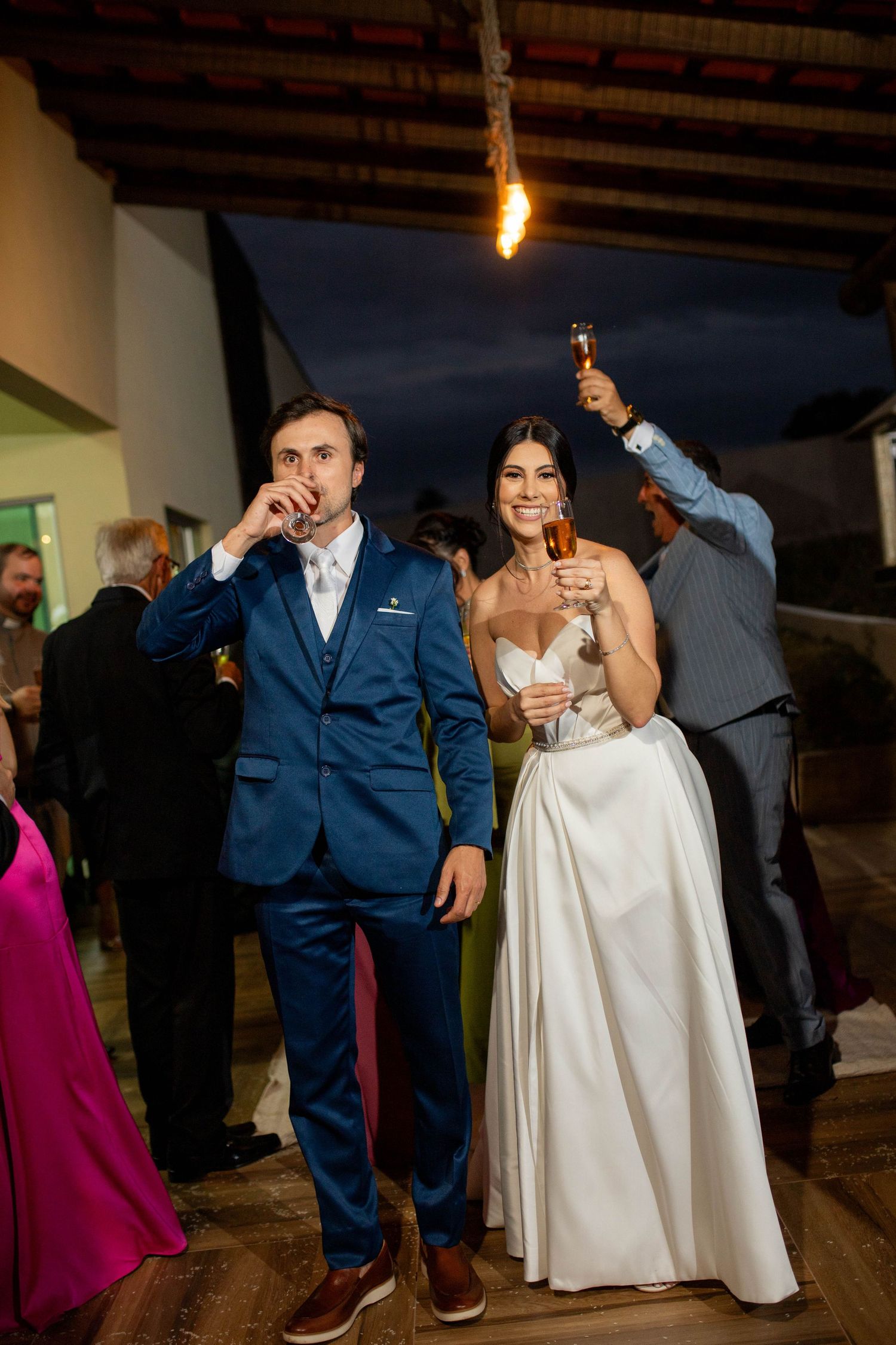Bride in white gown and groom in navy suit toasting drinks at outdoor wedding reception at night.