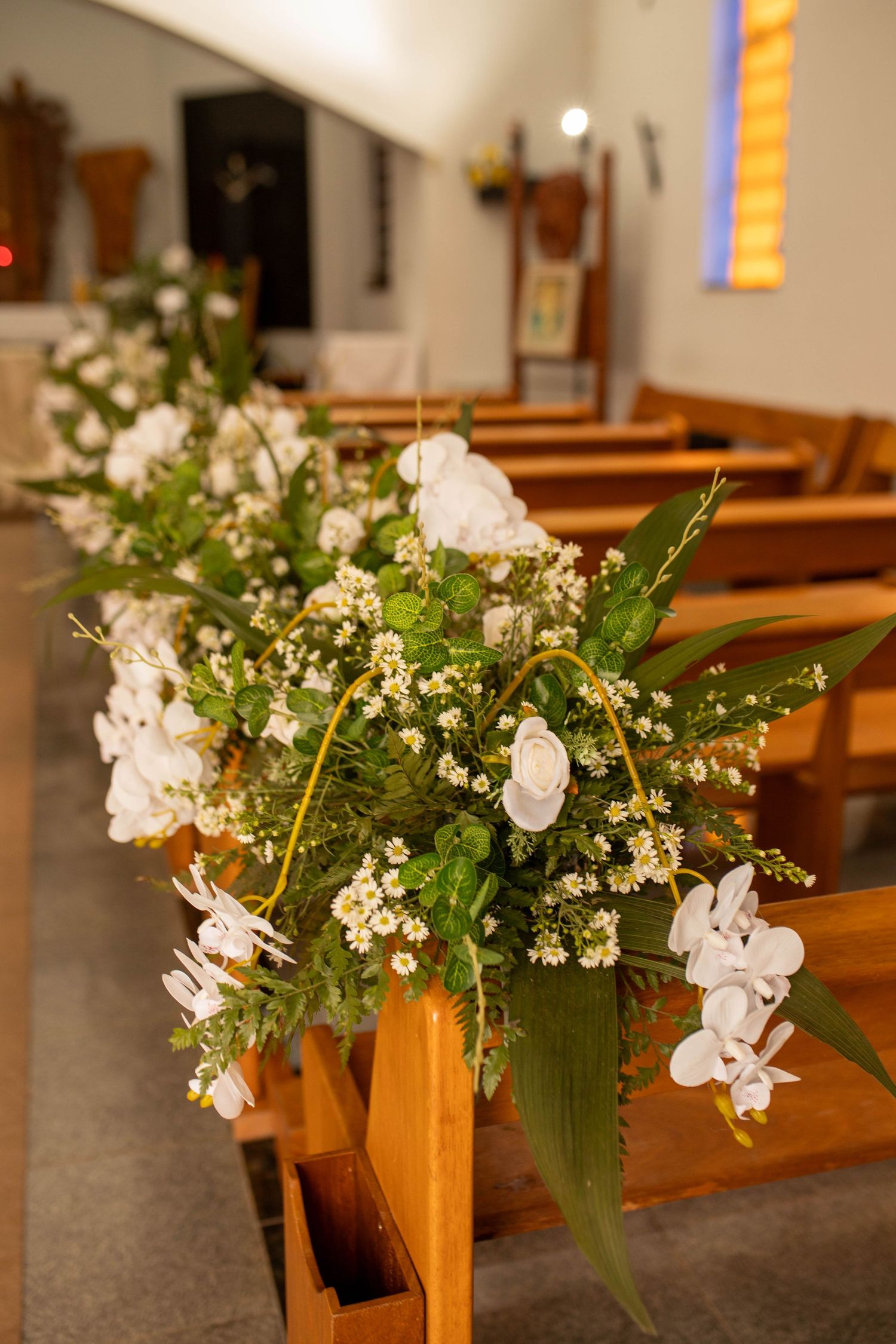 White orchids and wildflowers adorn wooden church pews, creating elegant wedding aisle decorations in a sunlit chapel.