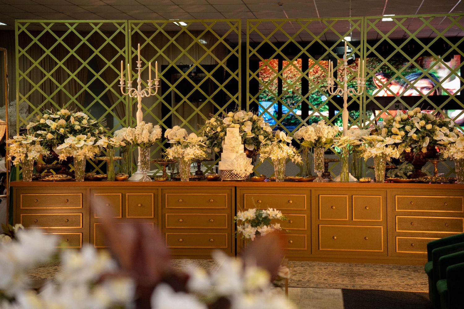 Elegant wedding reception dessert table with white floral arrangements, tiered cake, and gold wooden buffet in a decorated venue.