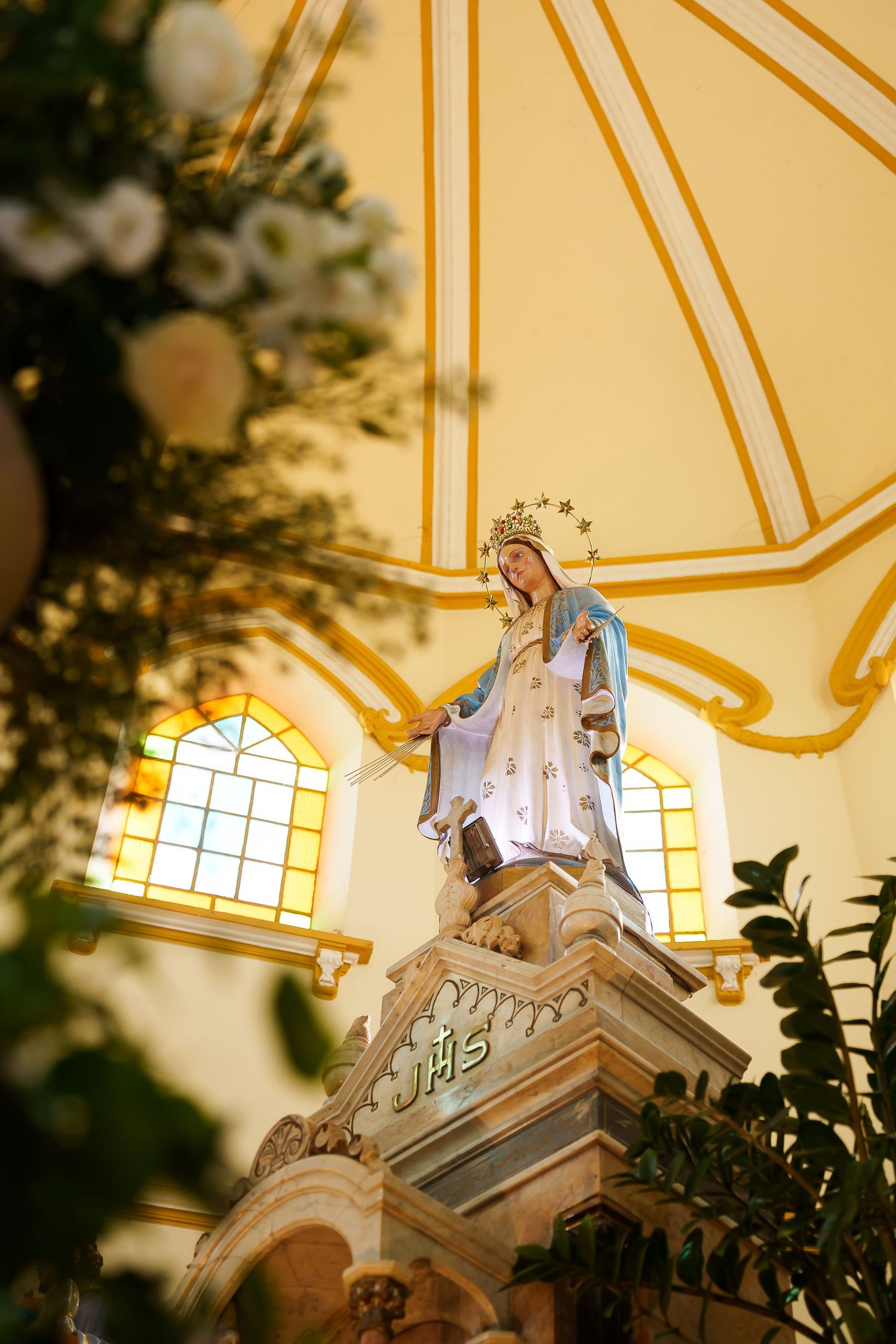 Statue of Virgin Mary with crown and blue robe on ornate pedestal inside a Catholic church with yellow domed ceiling.