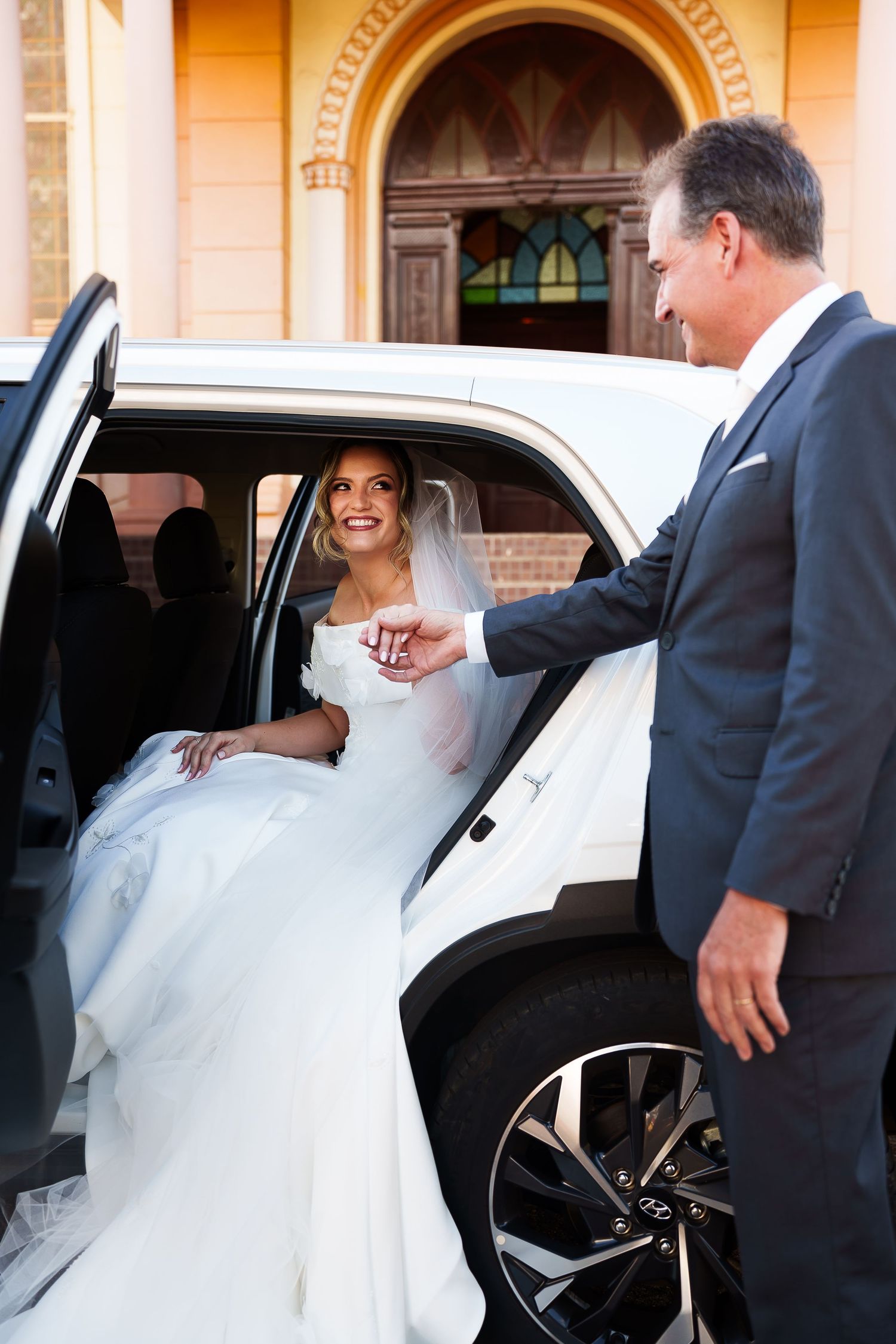Bride in white gown stepping out of white limousine, assisted by man in navy suit, outside church.