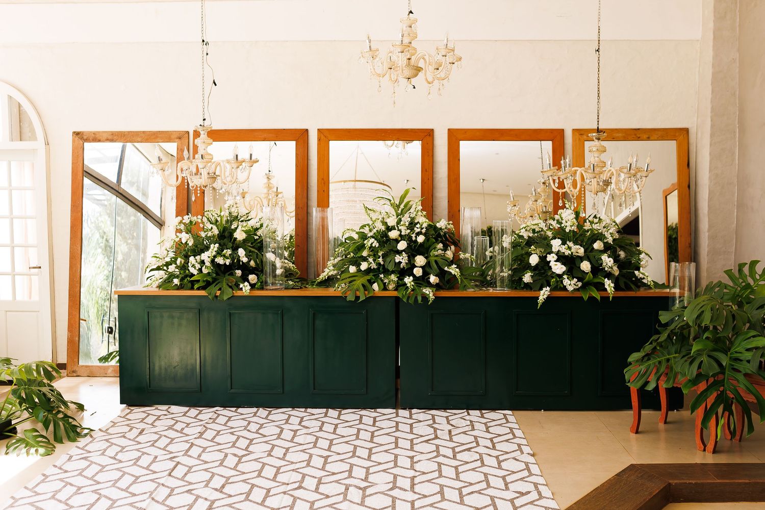 Elegant wedding reception desk with white floral arrangements, gold mirrors, and crystal chandeliers on geometric tile floor.