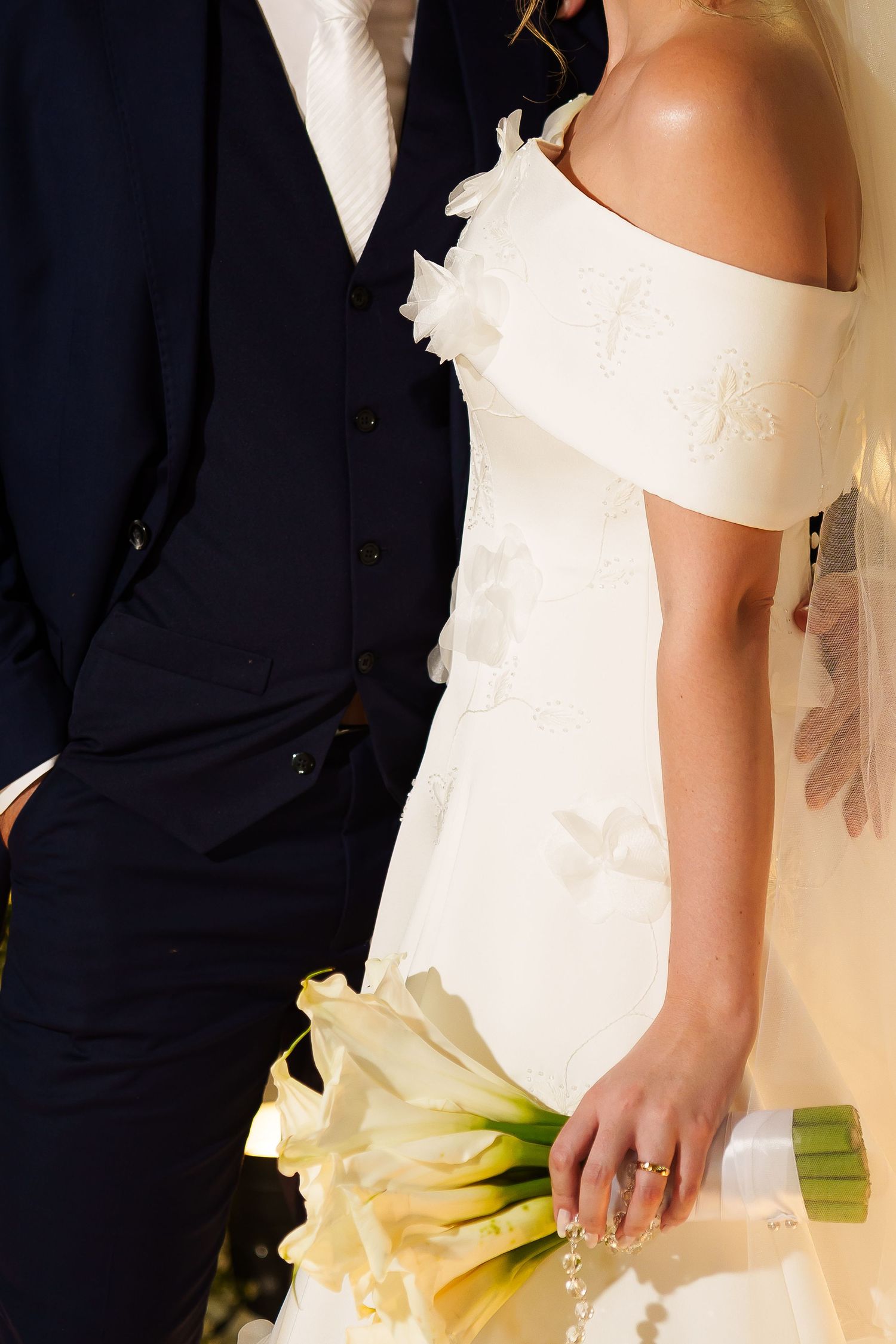 Bride in off-shoulder white floral gown holding calla lily bouquet stands beside groom in navy suit at wedding.