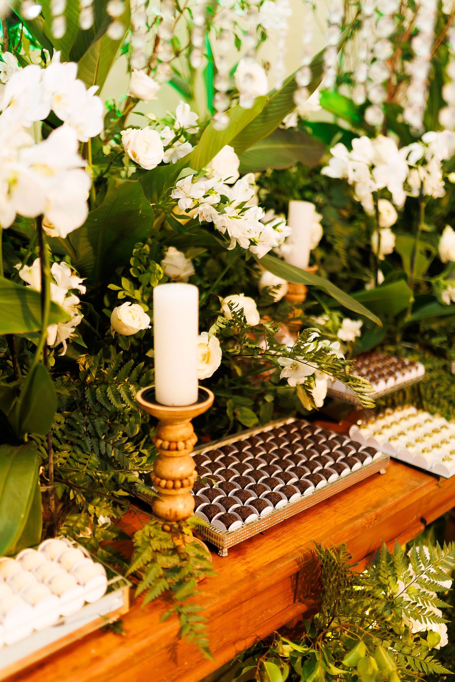 Elegant wedding dessert table with white candle, chocolate truffles, white flowers, and lush green foliage decor.