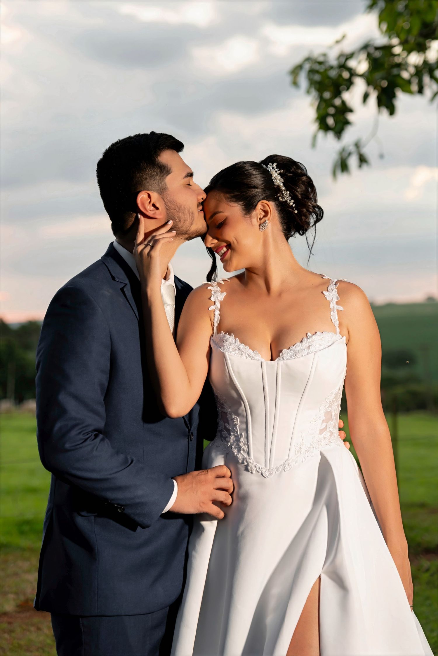 Groom in navy suit kisses bride in white lace wedding gown outdoors with green fields in background.