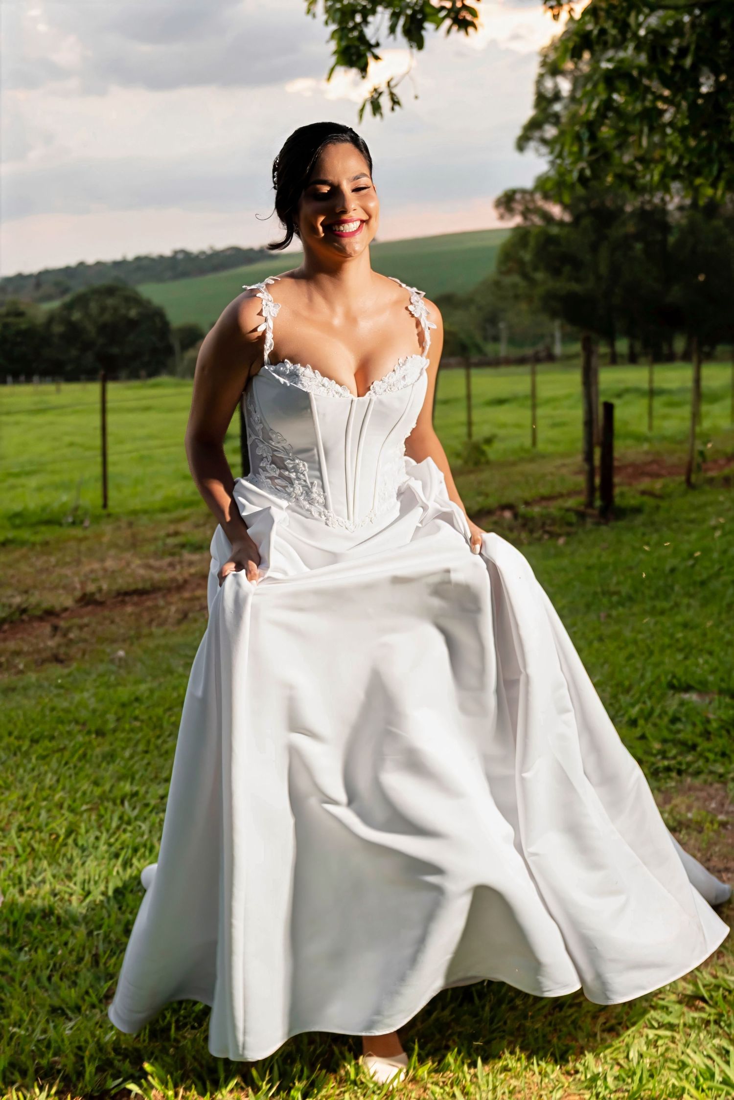 Smiling bride in white corset wedding gown twirling on green grass with rolling hills in background.