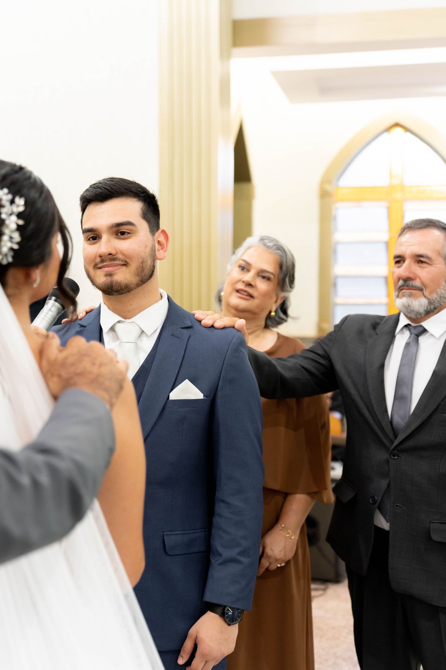 Groom in navy suit smiles during wedding ceremony as older couple places hands on his shoulders in church.