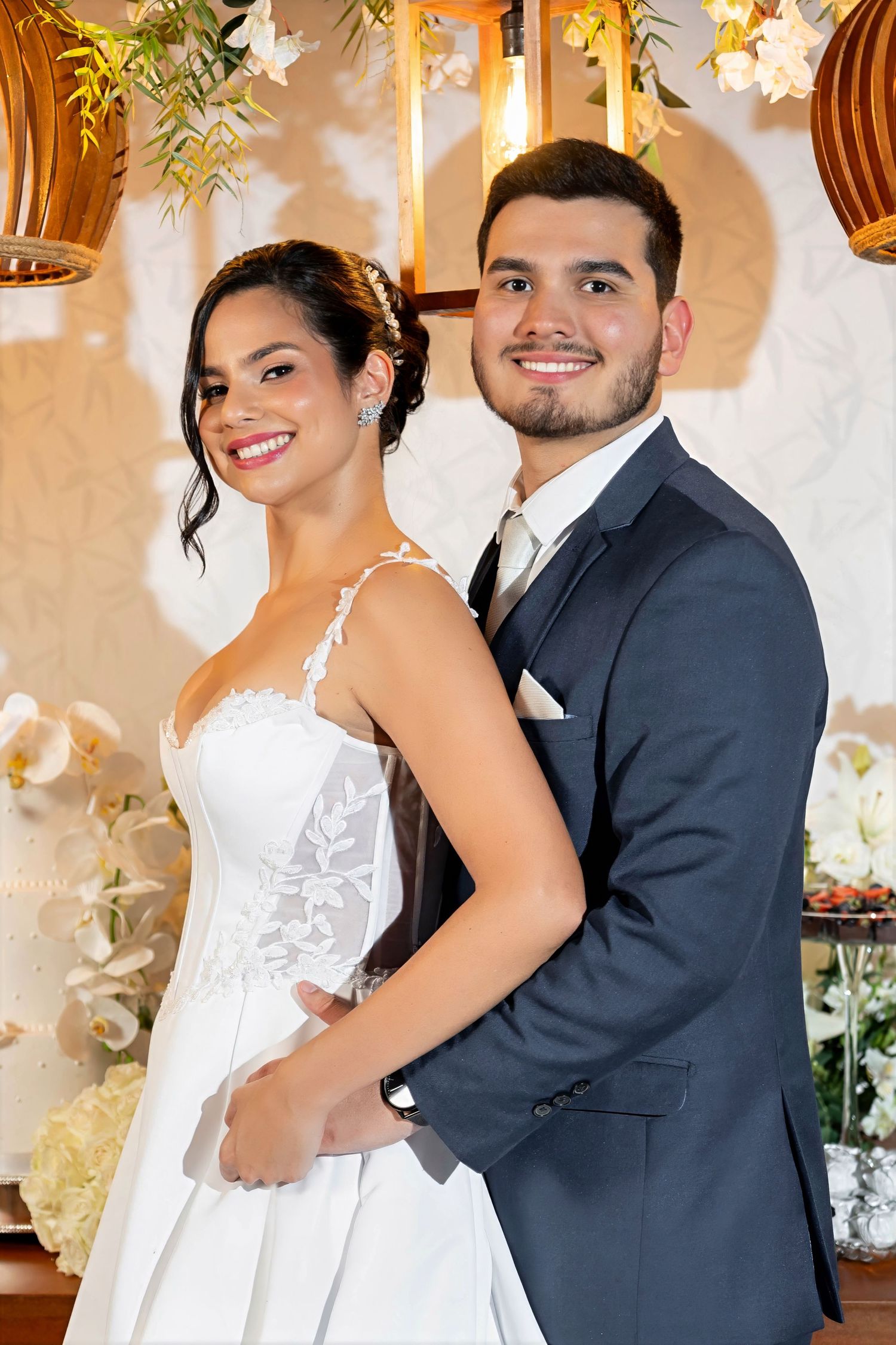 A smiling bride in a white lace gown and groom in a navy suit pose together at their elegant wedding reception.