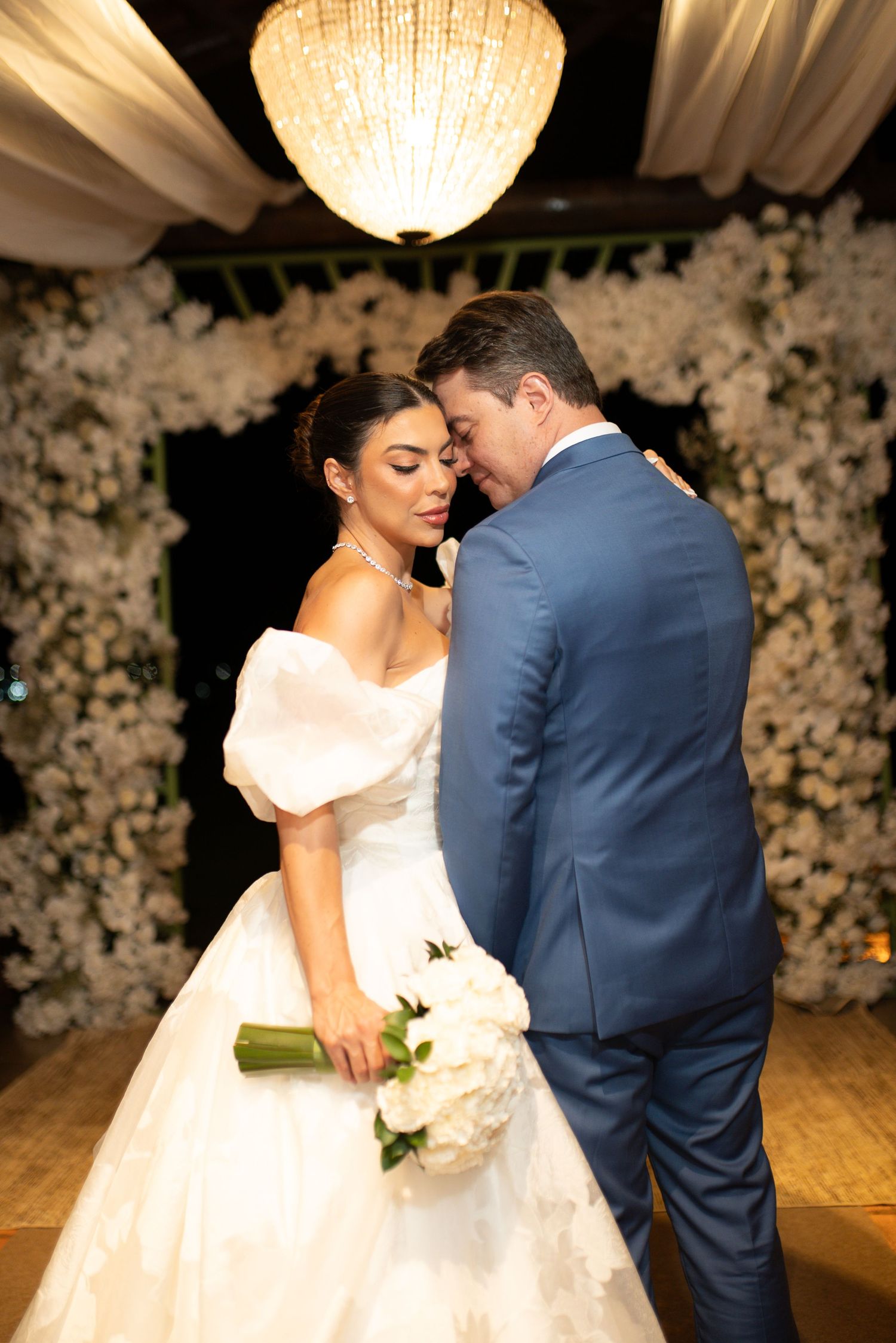 Bride in white off-shoulder gown holds bouquet while groom in blue suit leans in tenderly at floral wedding ceremony.