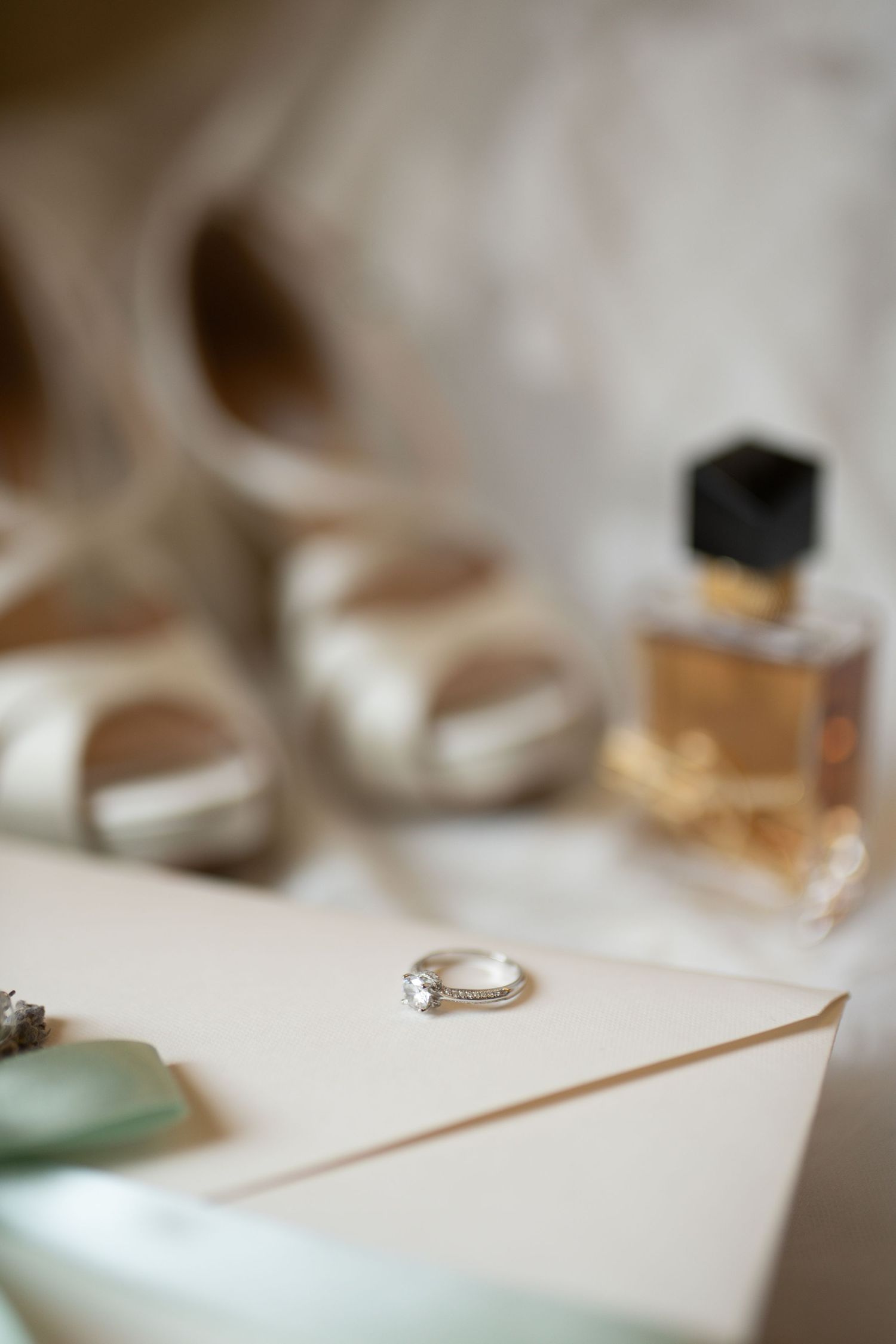 A diamond engagement ring rests on an envelope surrounded by bridal accessories including heels and perfume.