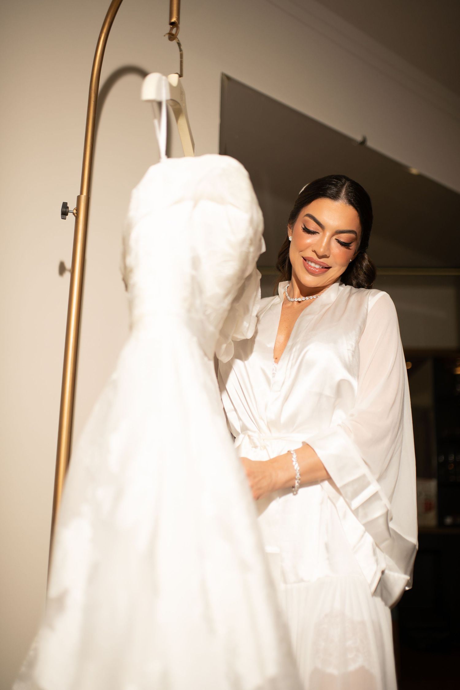 Bride in white satin robe smiling at her wedding dress hanging on a gold rack in a warmly lit room.