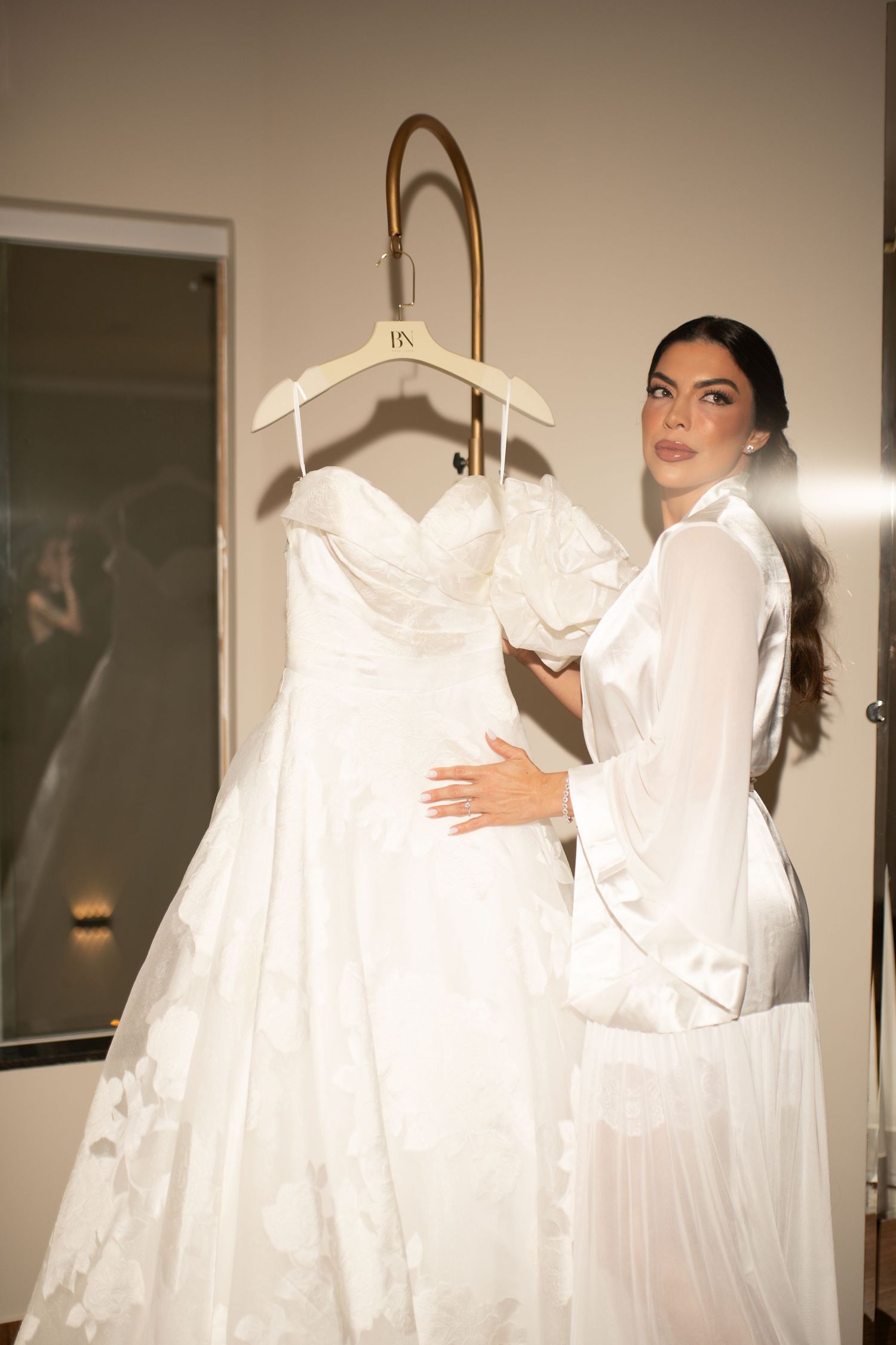 Bride in white satin robe admiring her lace wedding gown hanging on a gold hanger in a warmly lit room.