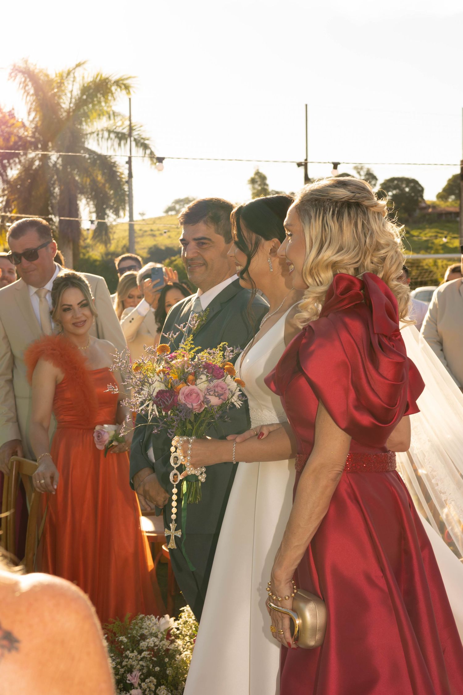 Bride in white gown receives kiss from woman in red dress during outdoor wedding ceremony at golden hour.