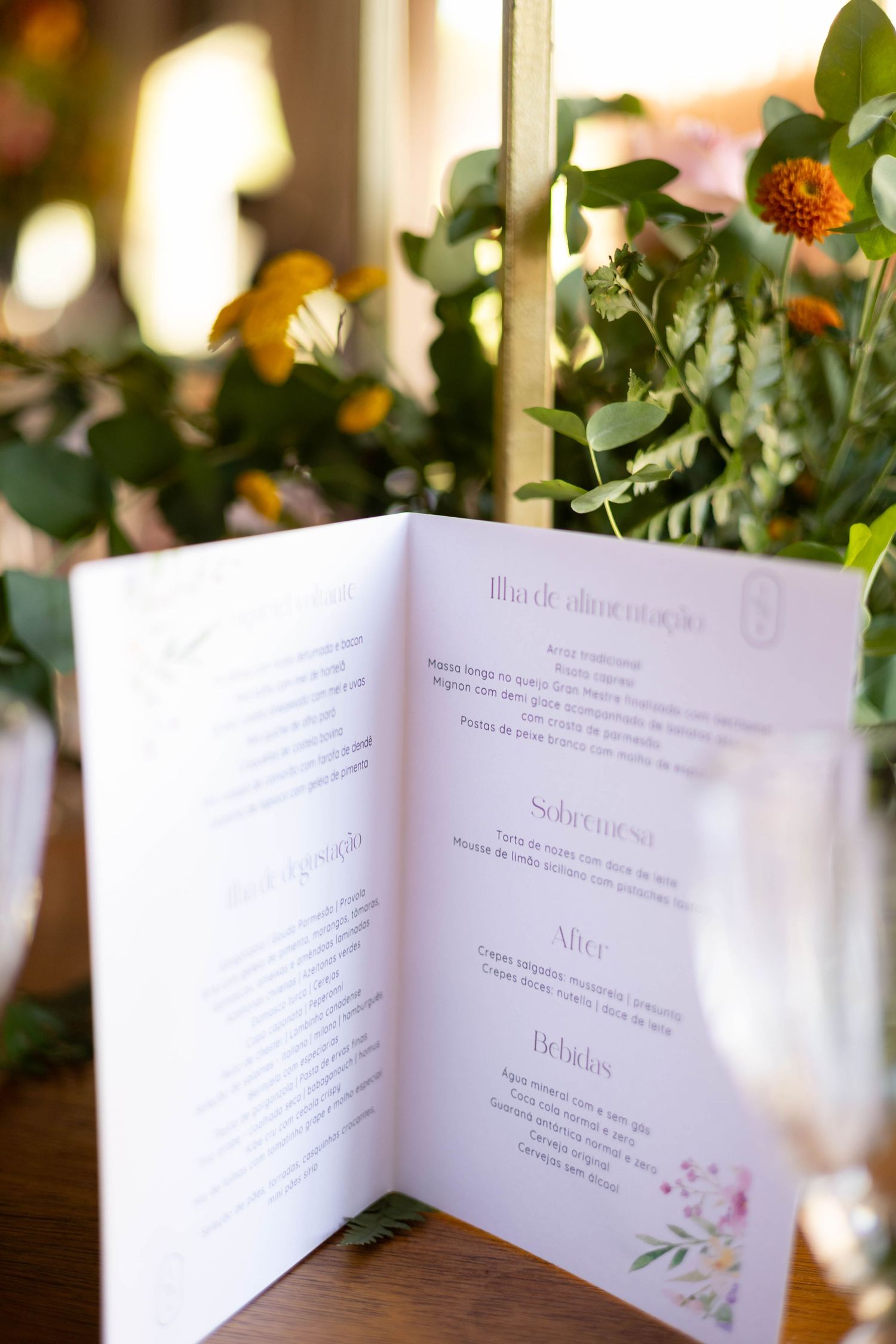 Elegant wedding menu card displayed on a wooden table surrounded by lush greenery and orange floral arrangements.