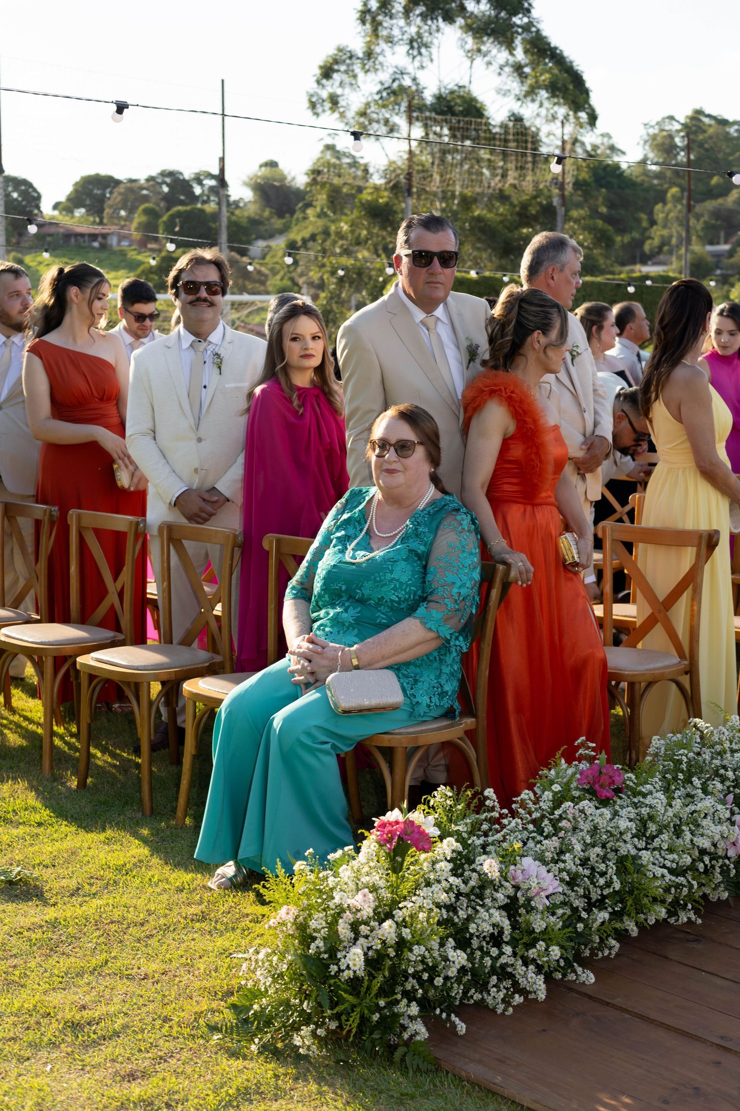 Outdoor wedding ceremony guests in colorful formal attire seated and standing on green lawn with floral arrangements.