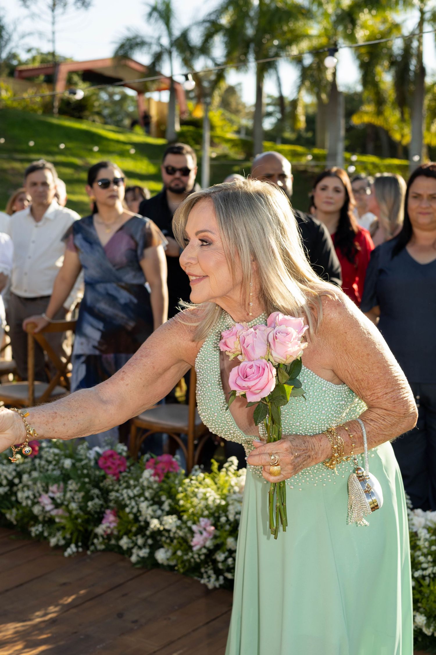 Elegant woman in mint green gown holding pink roses at an outdoor wedding ceremony with guests seated behind her.