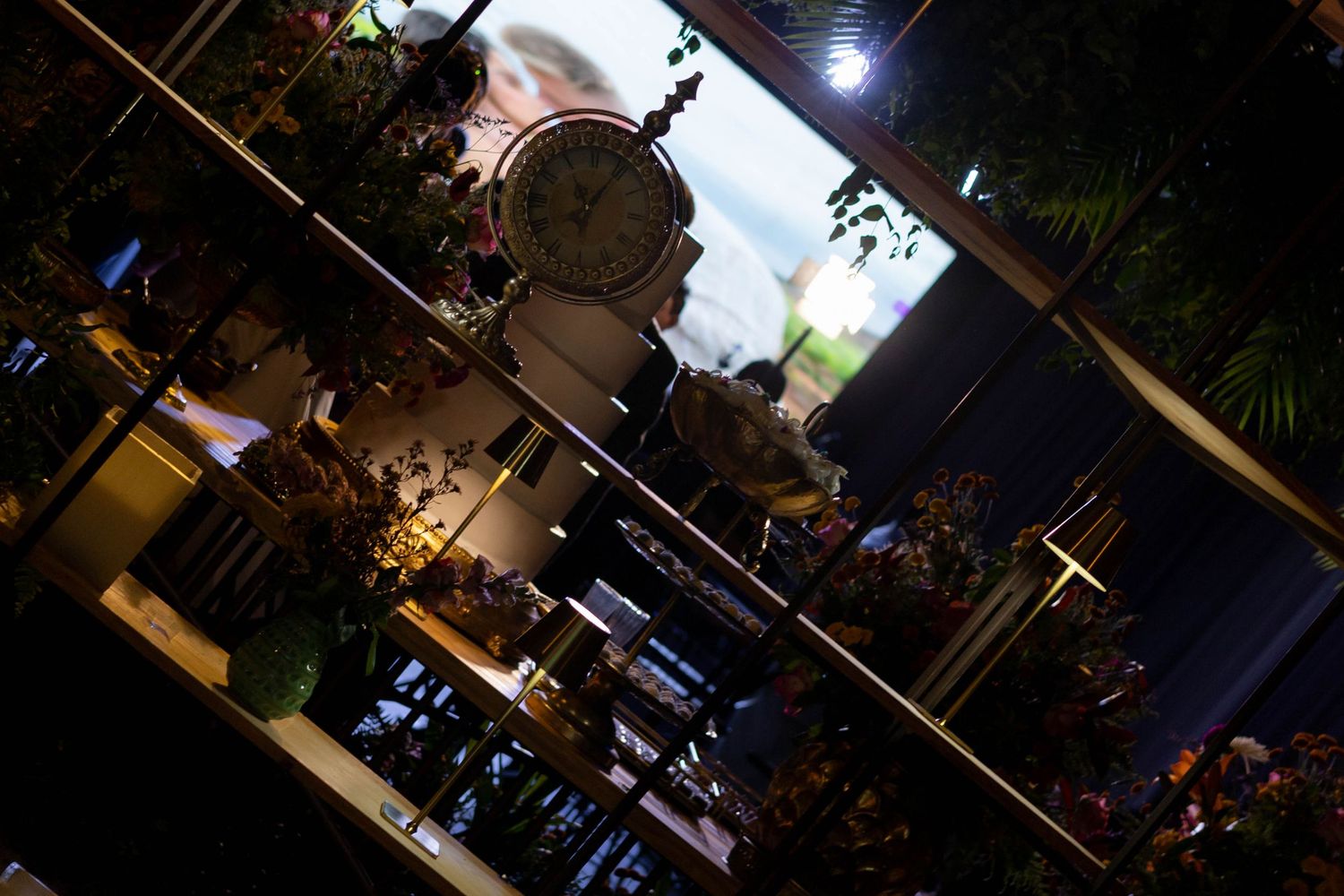 Dimly lit indoor display with a vintage double-sided clock surrounded by lush green plants on tiered shelves.