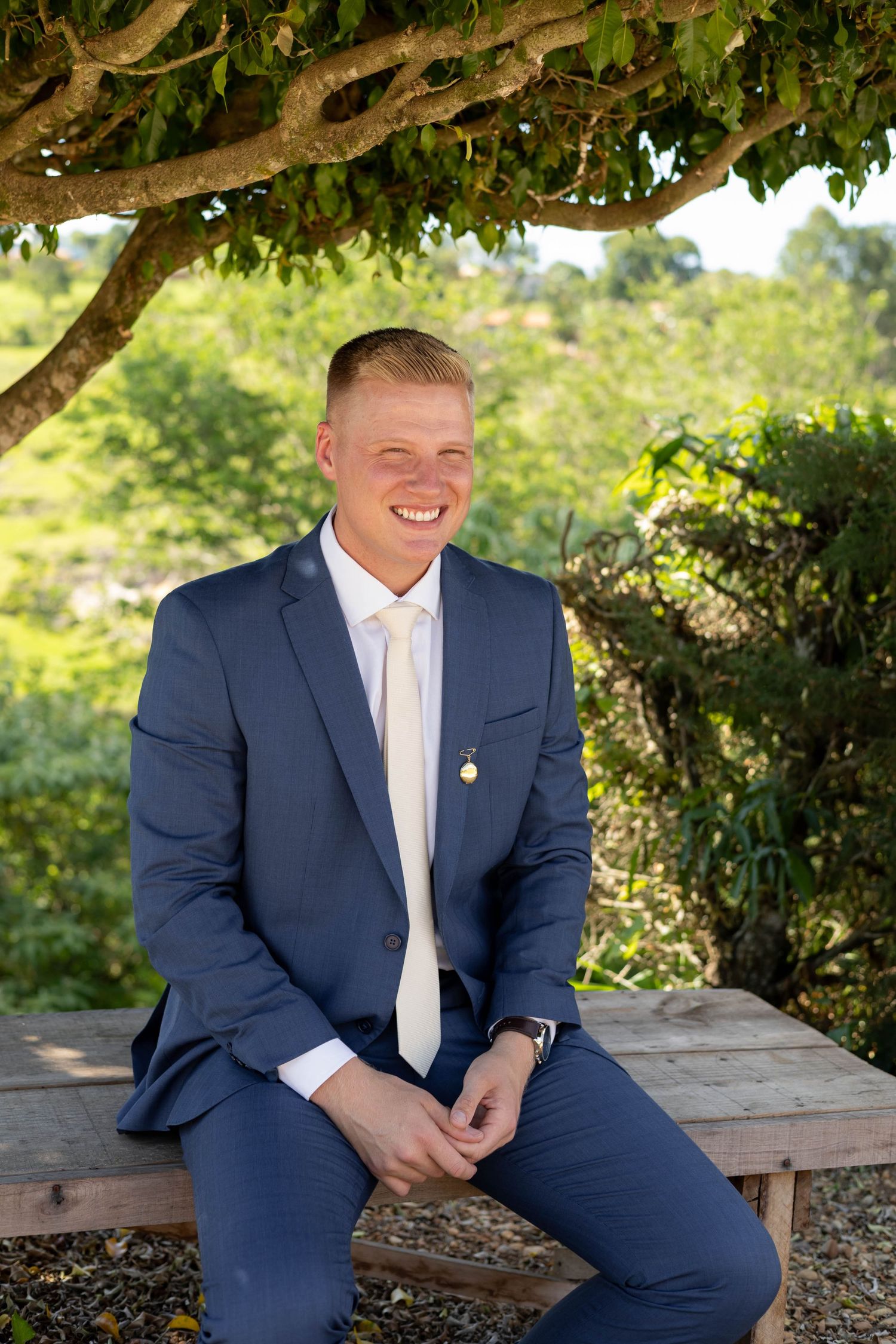 Smiling young man in navy blue suit and cream tie sitting outdoors under a tree with lush green background.