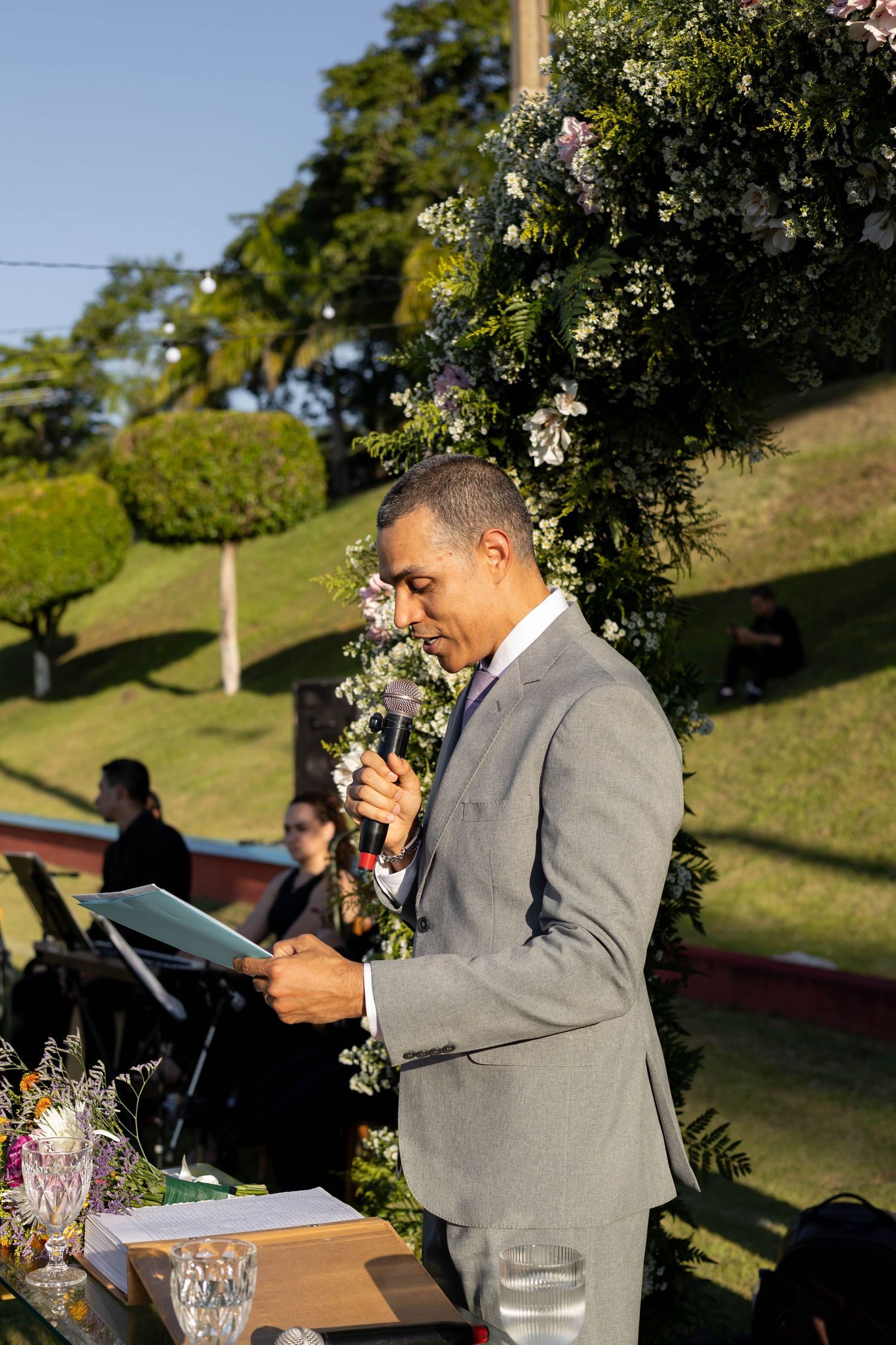 Man in gray suit speaking at outdoor wedding ceremony with floral arch backdrop.