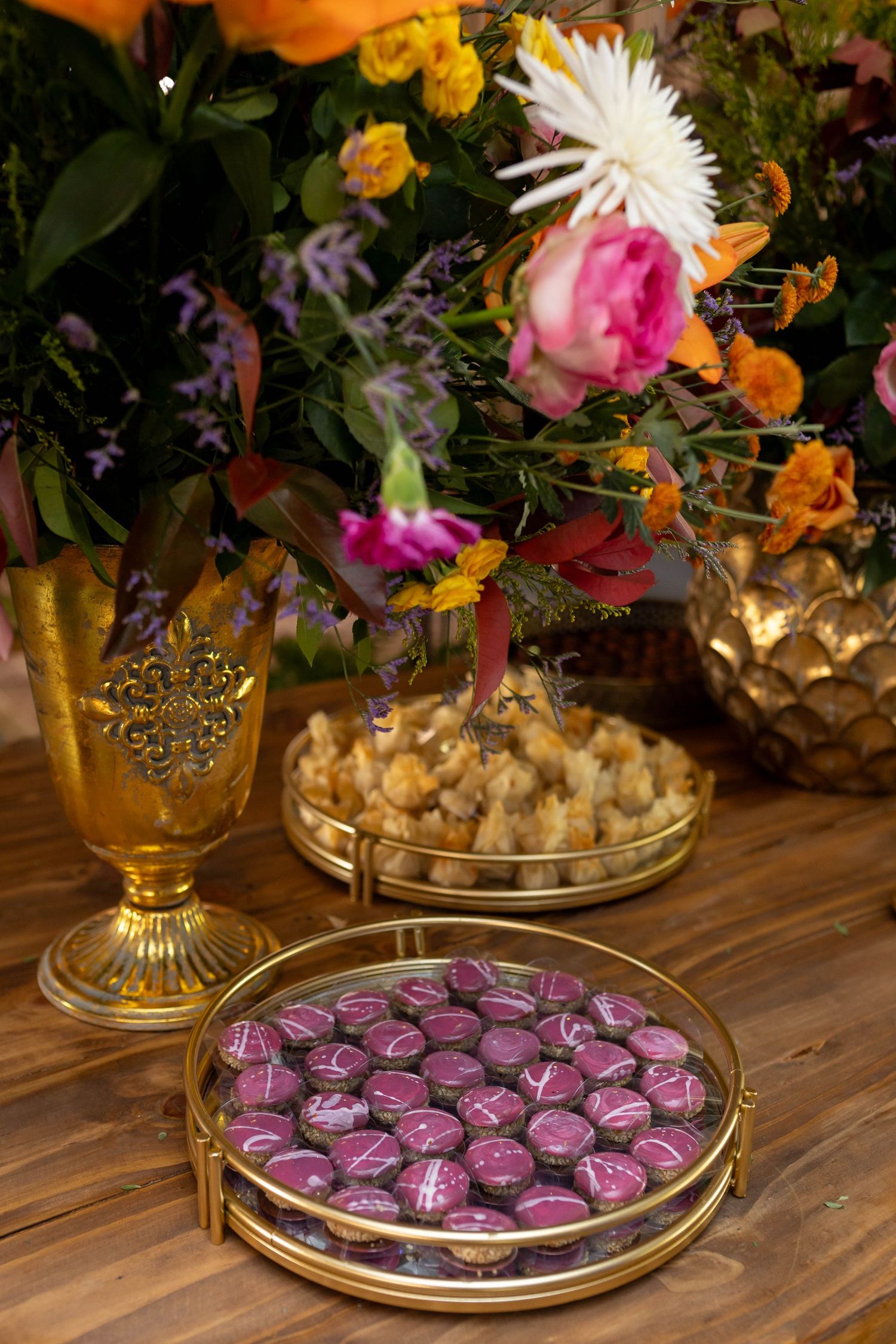 Gold tray with purple chocolate candies beside a floral arrangement with pink roses and orange blooms on a wooden table.