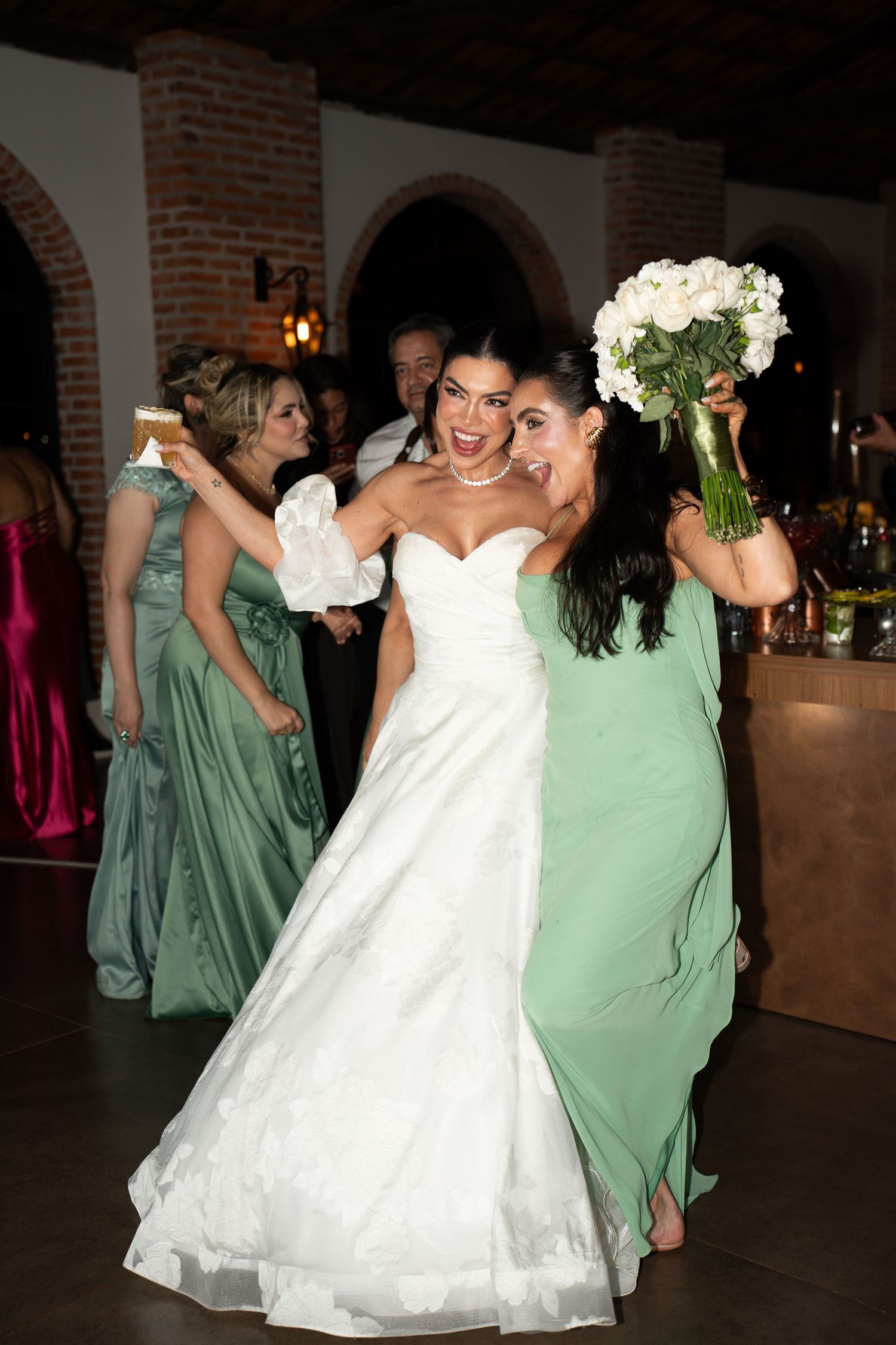 Bride in white gown holds bouquet with bridesmaid in sage green dress at elegant wedding reception.