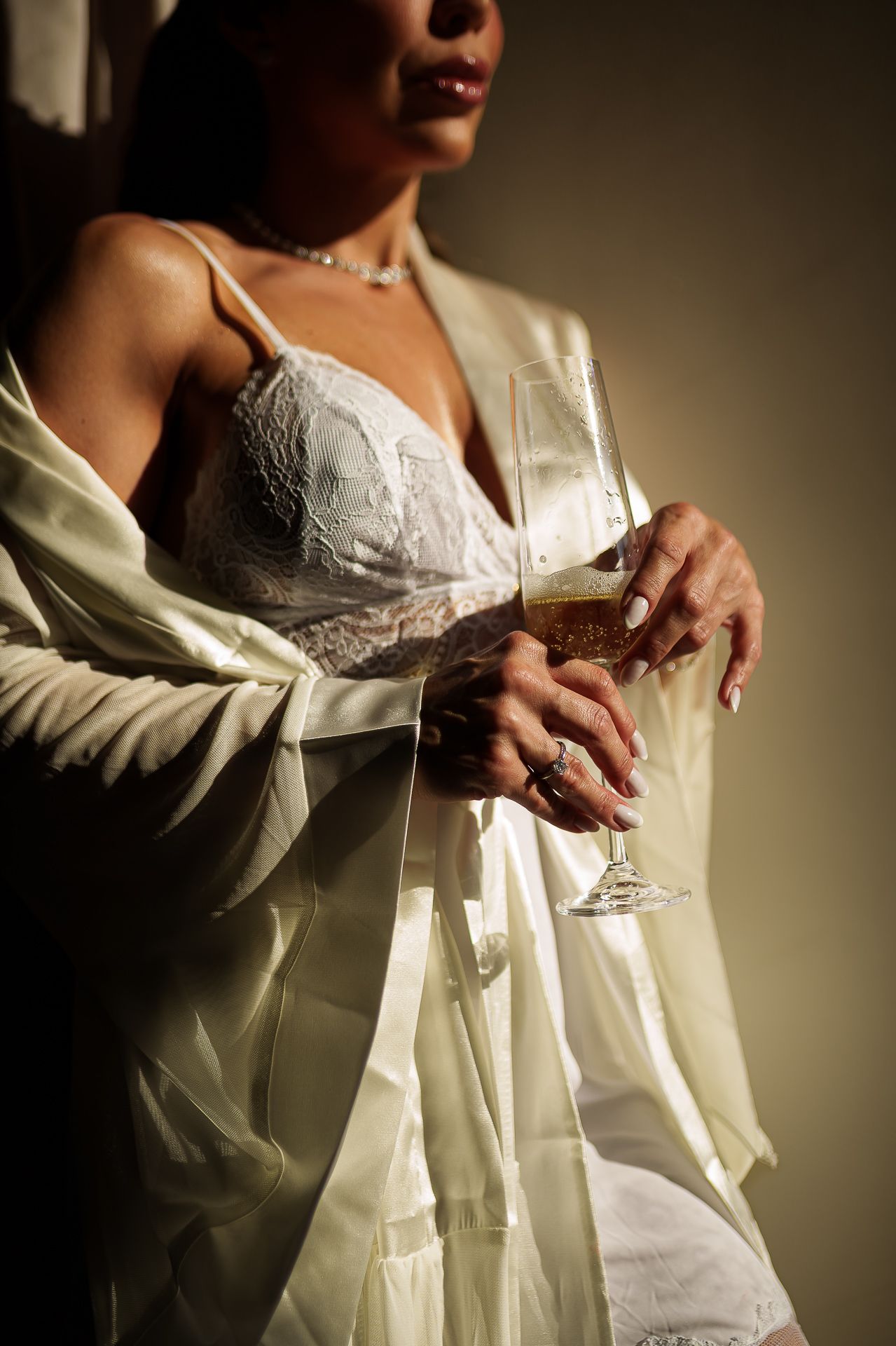 Woman in white lace lingerie and satin robe holding a champagne glass, adorned with jewelry.