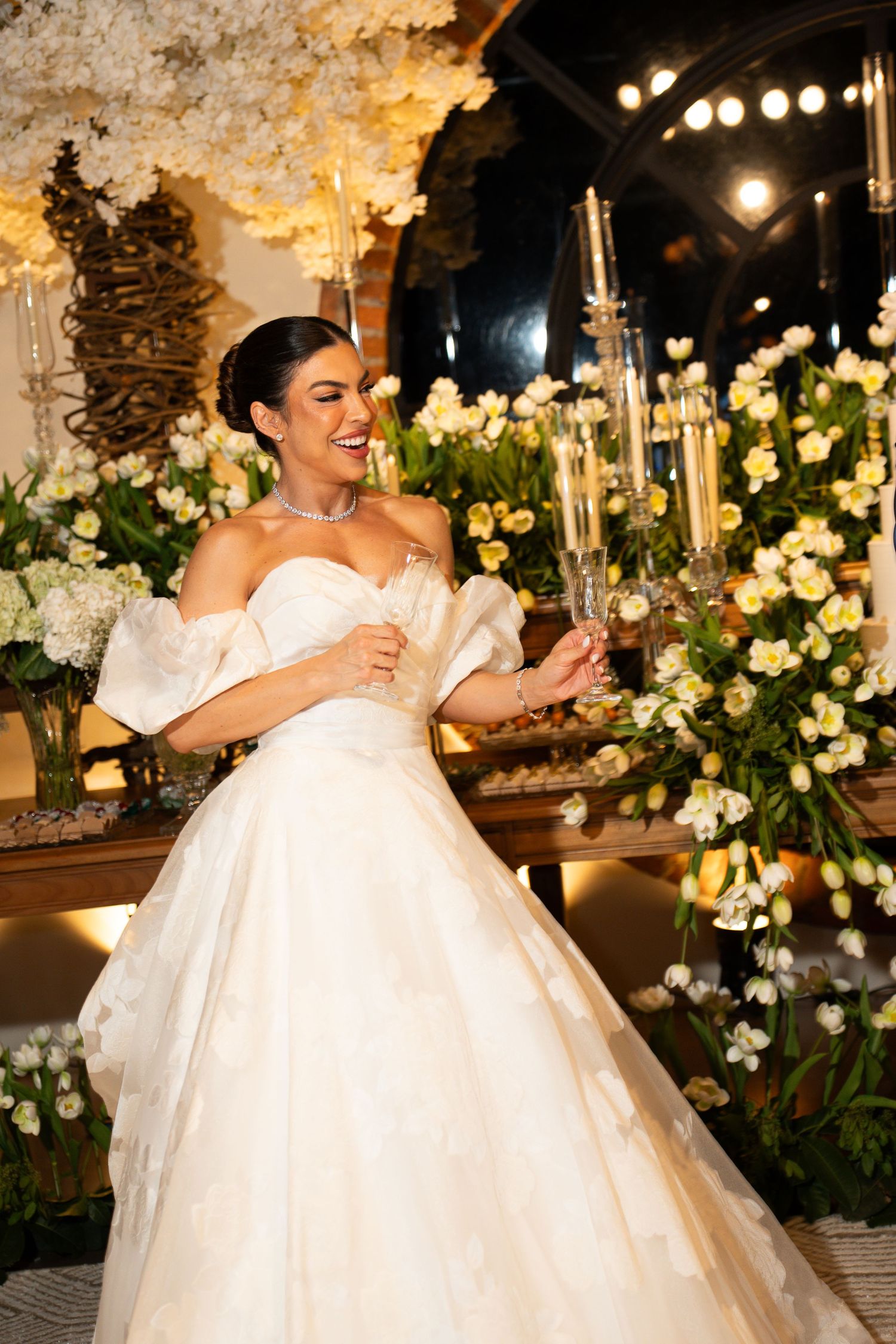 Bride in off-shoulder ball gown holding champagne flute, surrounded by white floral arrangements at elegant wedding reception.