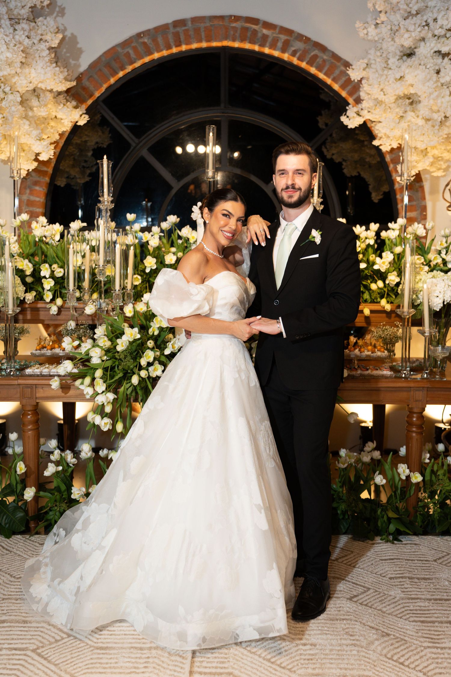Bride in white ball gown and groom in black tuxedo pose together at elegant floral wedding reception.