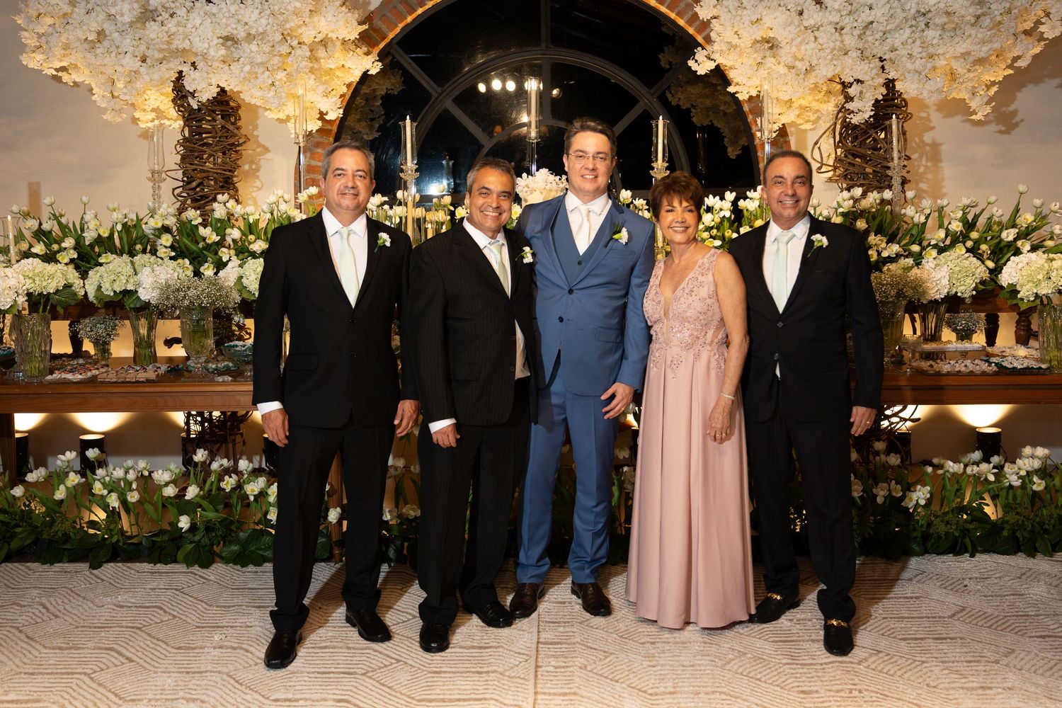 Five elegantly dressed guests pose together at a floral wedding reception with white flower arrangements.