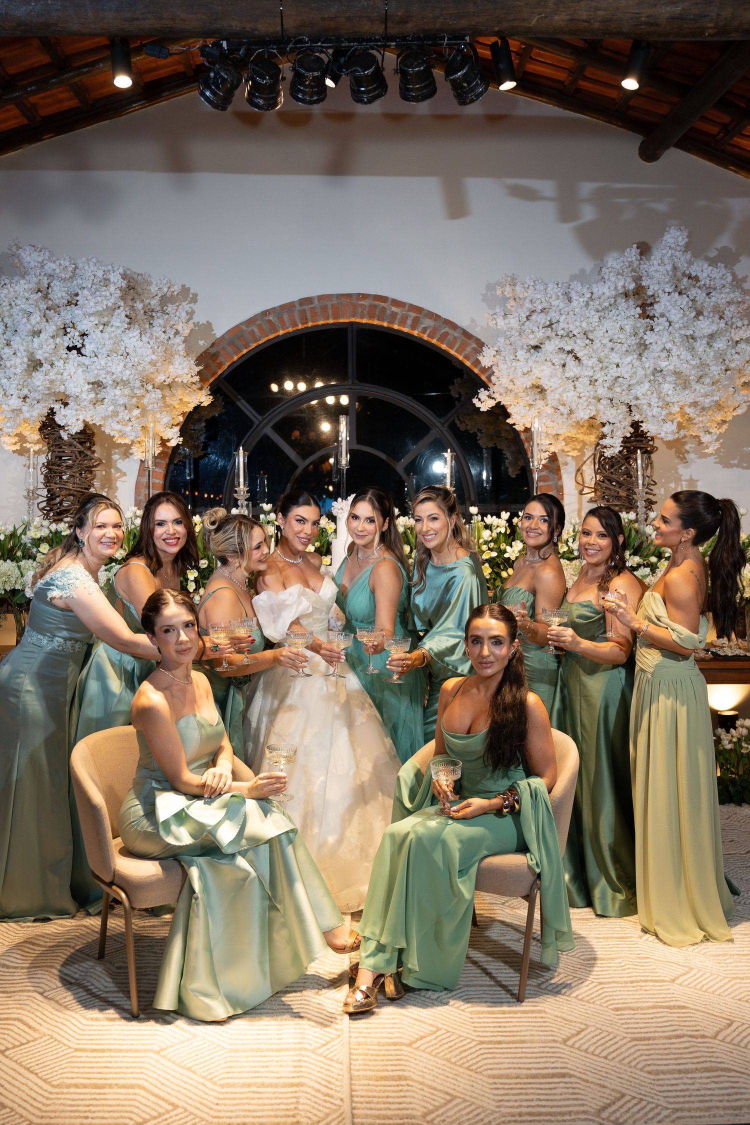 Bride in white gown poses with bridesmaids in sage green dresses, holding champagne glasses at elegant floral wedding reception.