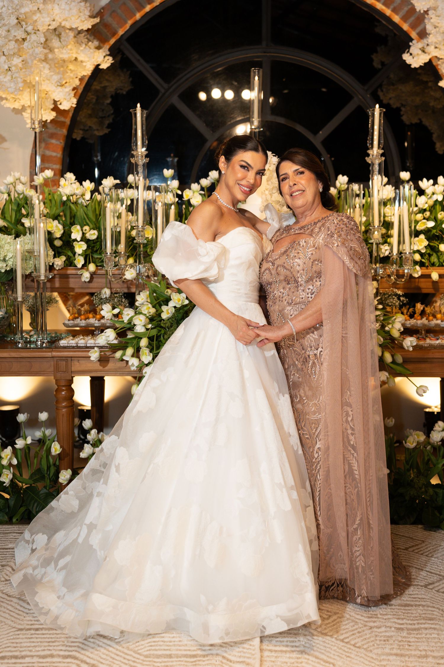 Bride in white ball gown poses with older woman in beige lace gown at elegant floral wedding reception.