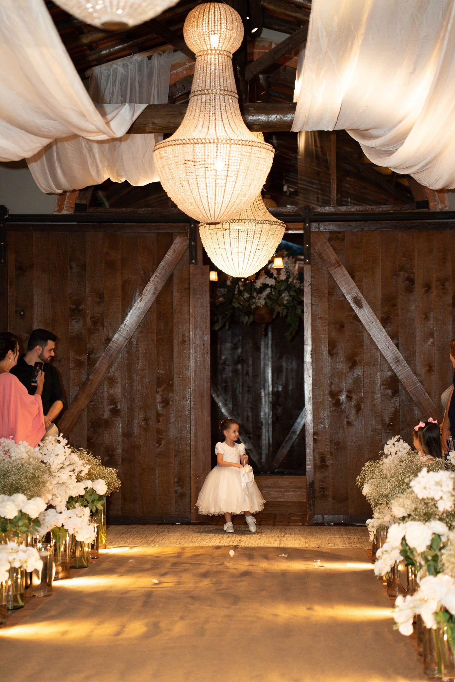Flower girl walks down barn wedding aisle lined with white floral arrangements under elegant chandelier.