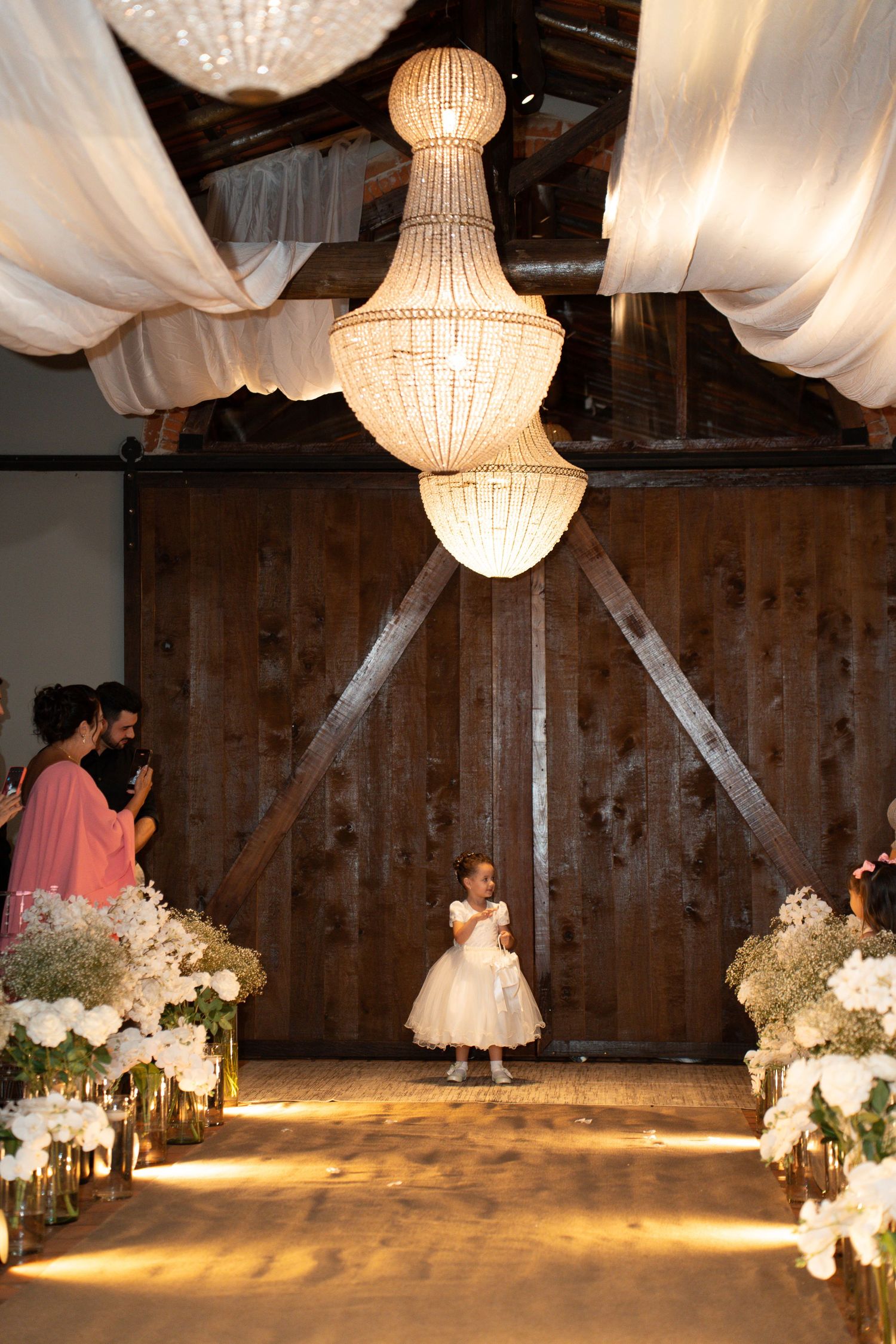 A flower girl in a white dress stands at a barn wedding aisle adorned with floral arrangements under a crystal chandelier.