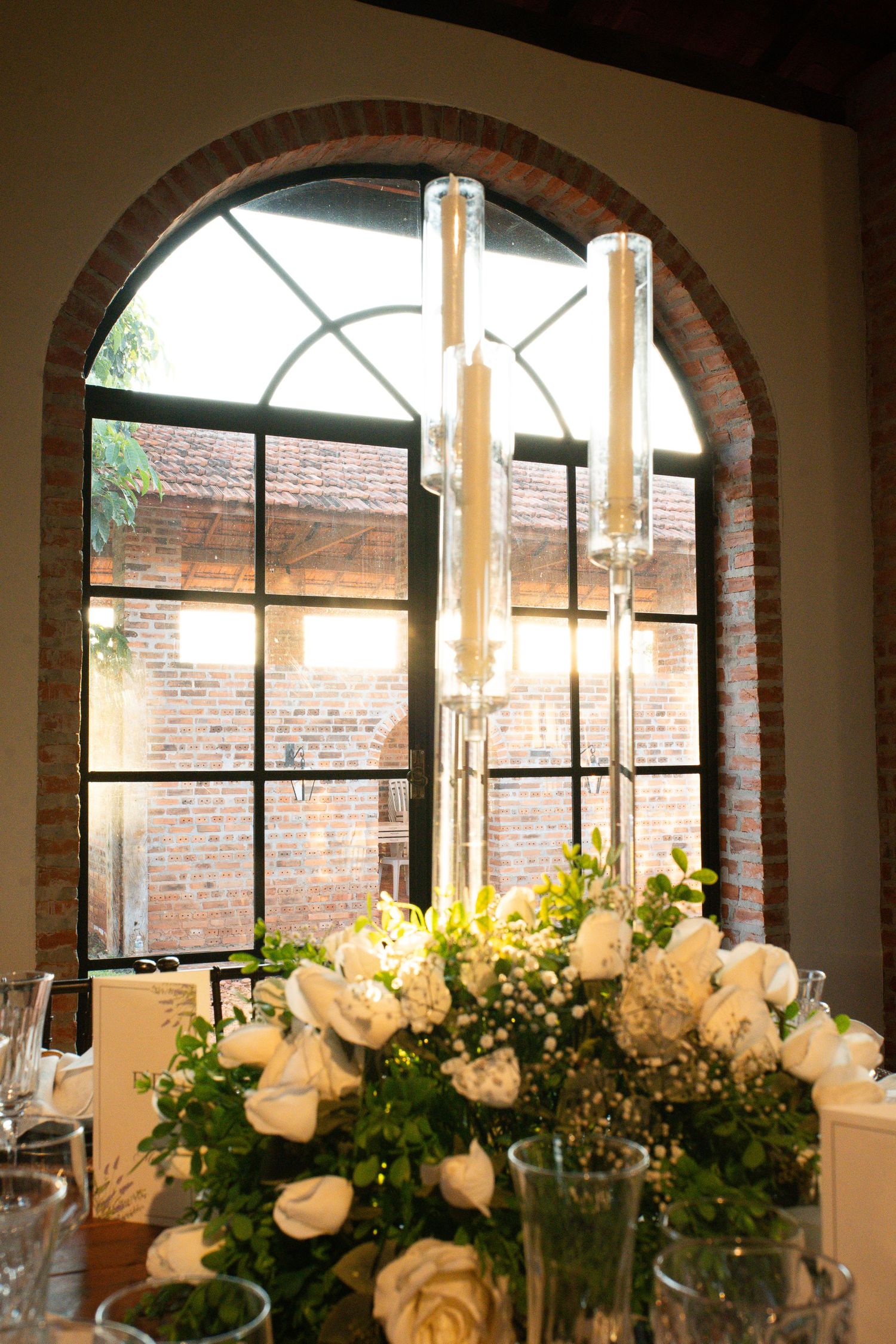 White roses and greenery centerpiece on a wedding reception table with tall glass candle holders and arched brick window.