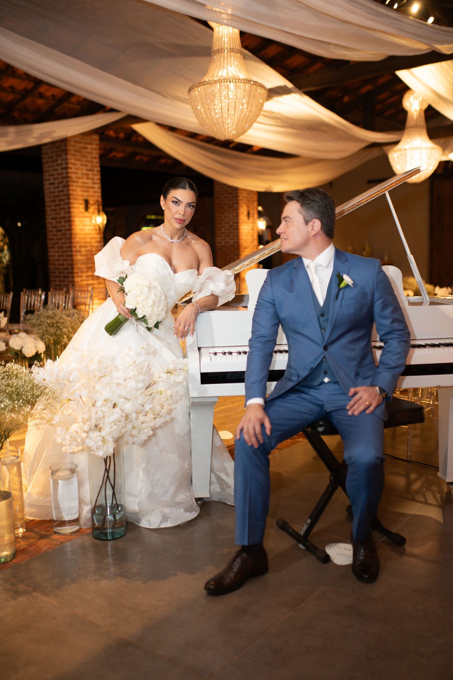 Bride in white gown holding bouquet leans on white piano beside groom in blue suit at elegant wedding reception.