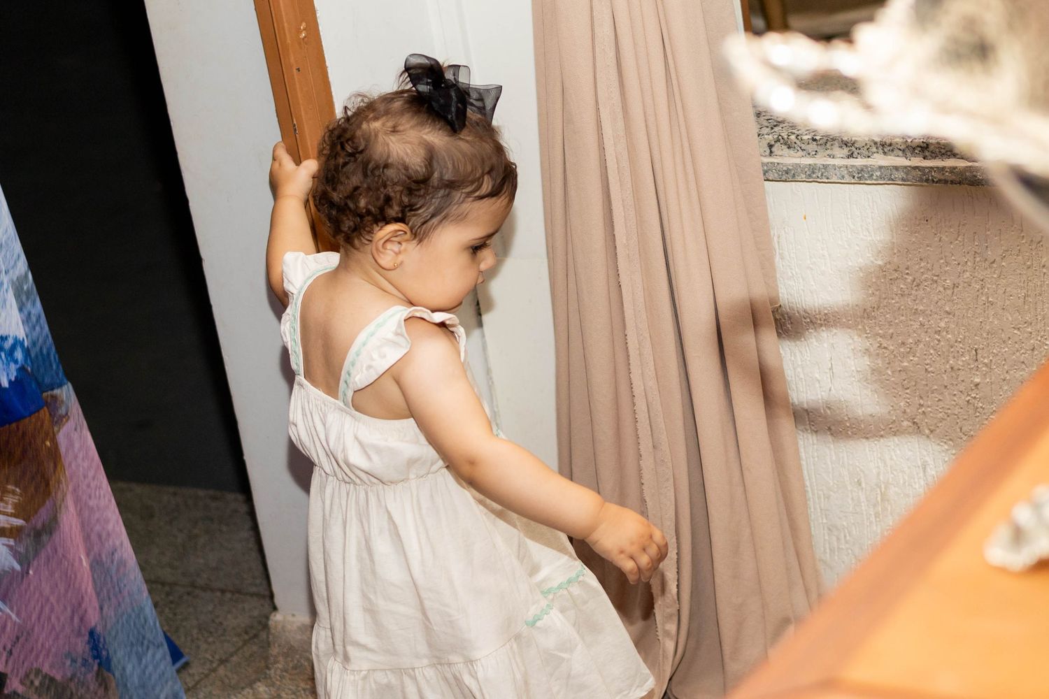Toddler girl in white dress exploring indoors, reaching toward a sheer curtain near a doorway.