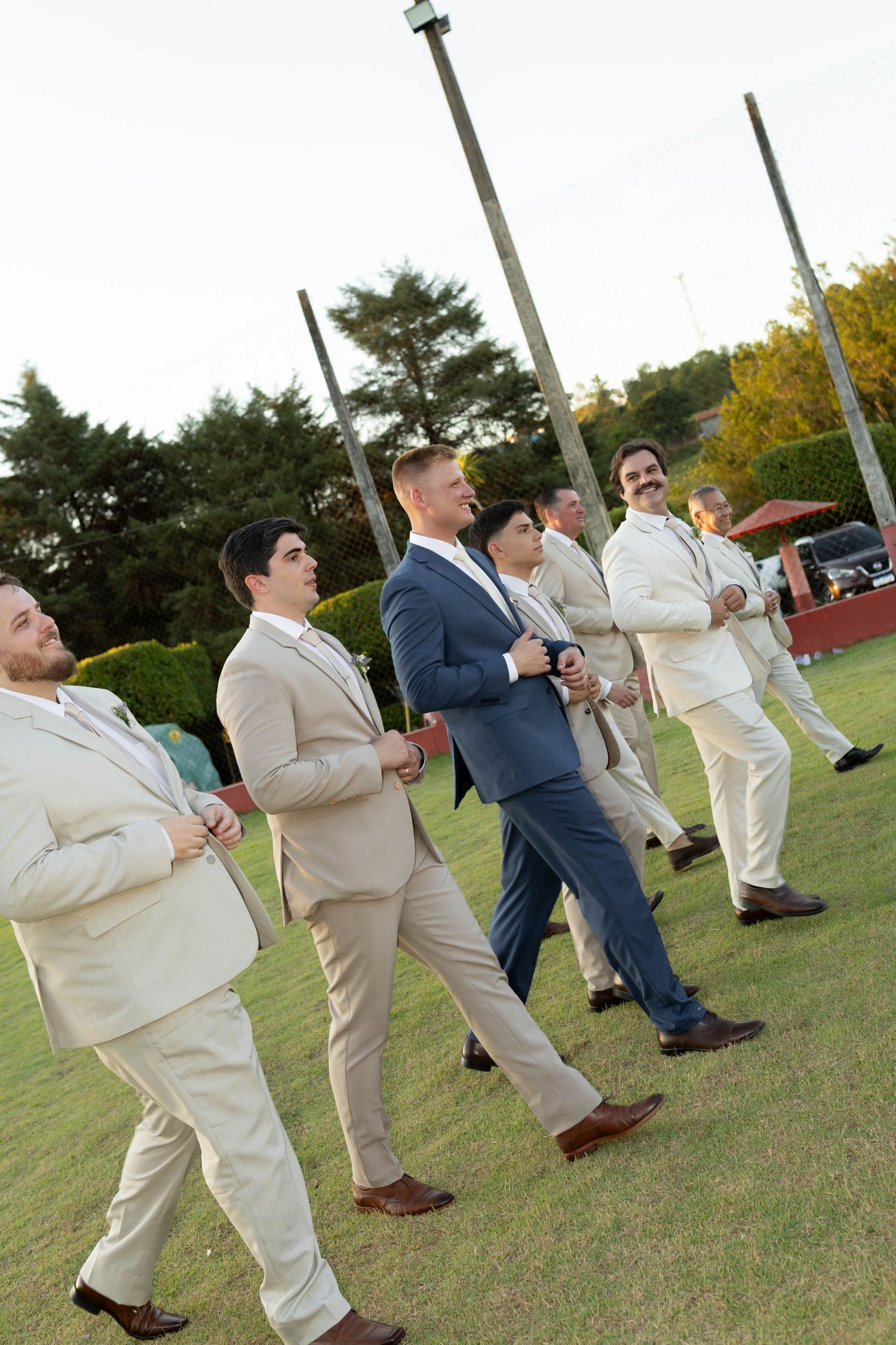Group of men in suits walking outdoors on grass at a formal event, wearing beige and blue attire.
