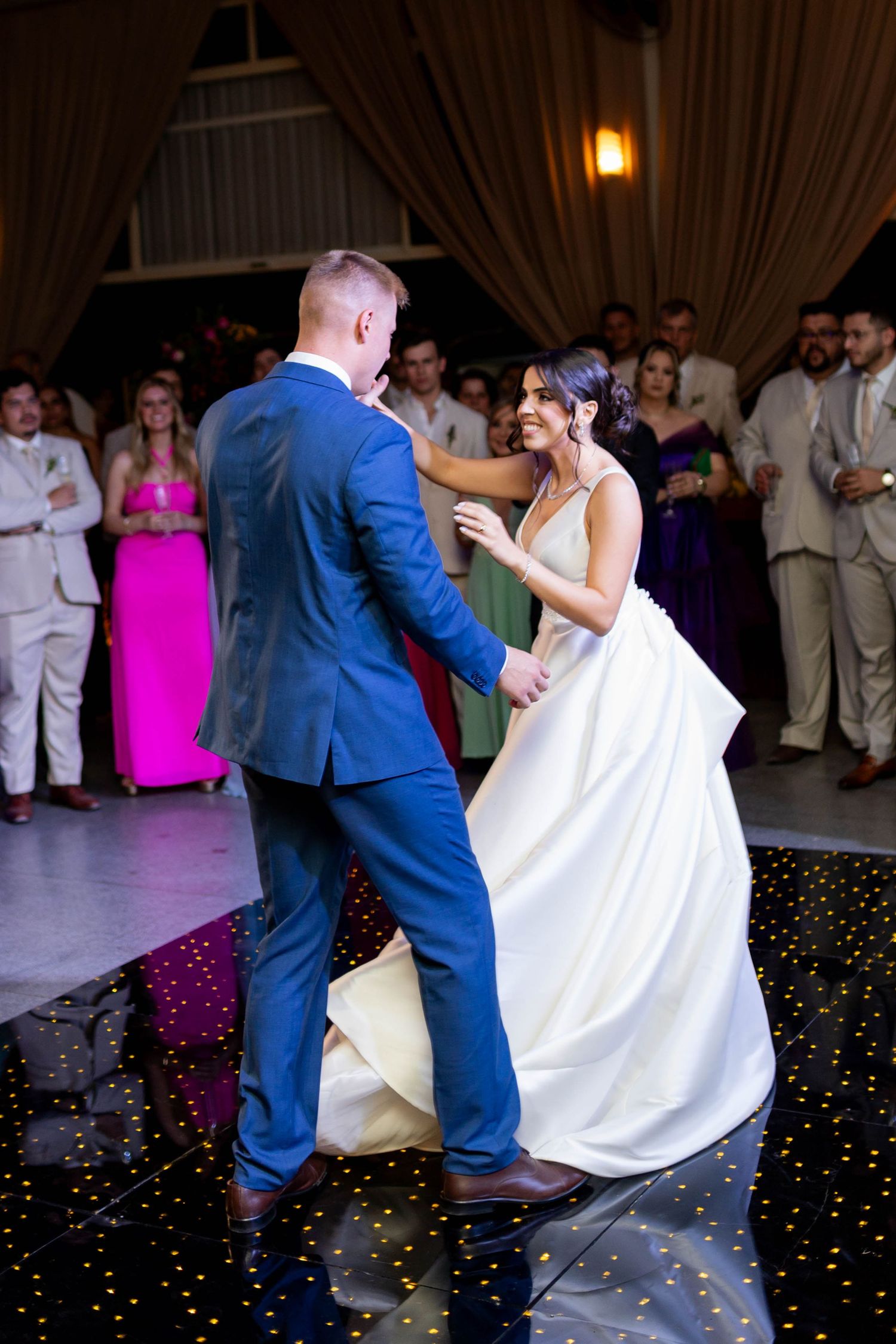 Bride and groom share their first dance on a starlit floor while wedding guests watch in a elegant draped venue.