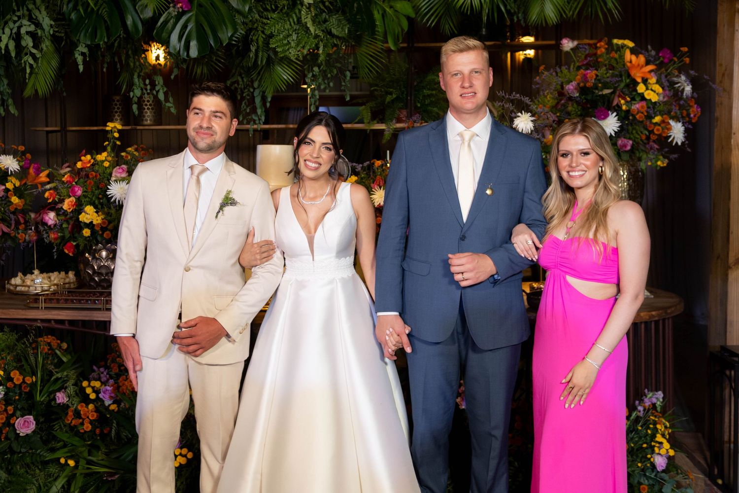 Two couples pose together at a wedding, bride in white gown, groom in tan suit, guests in blue suit and pink dress.