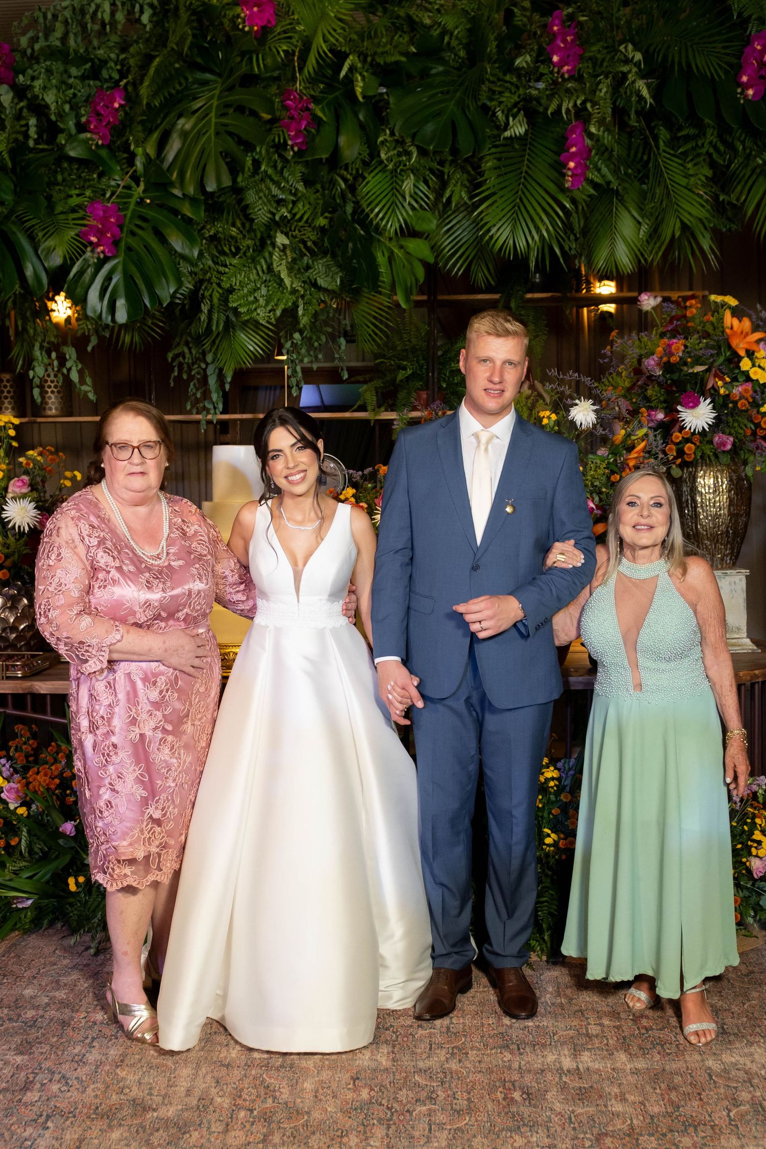 Wedding couple in formal attire poses with two women in pink and green dresses before a lush floral backdrop.