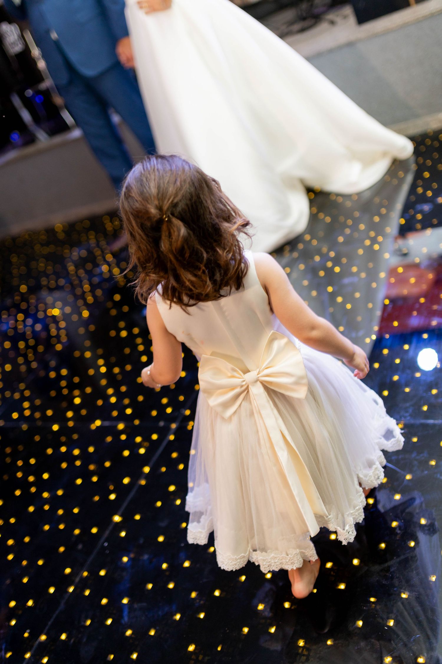Young flower girl in white dress with gold bow dances on sparkling LED dance floor at wedding reception.
