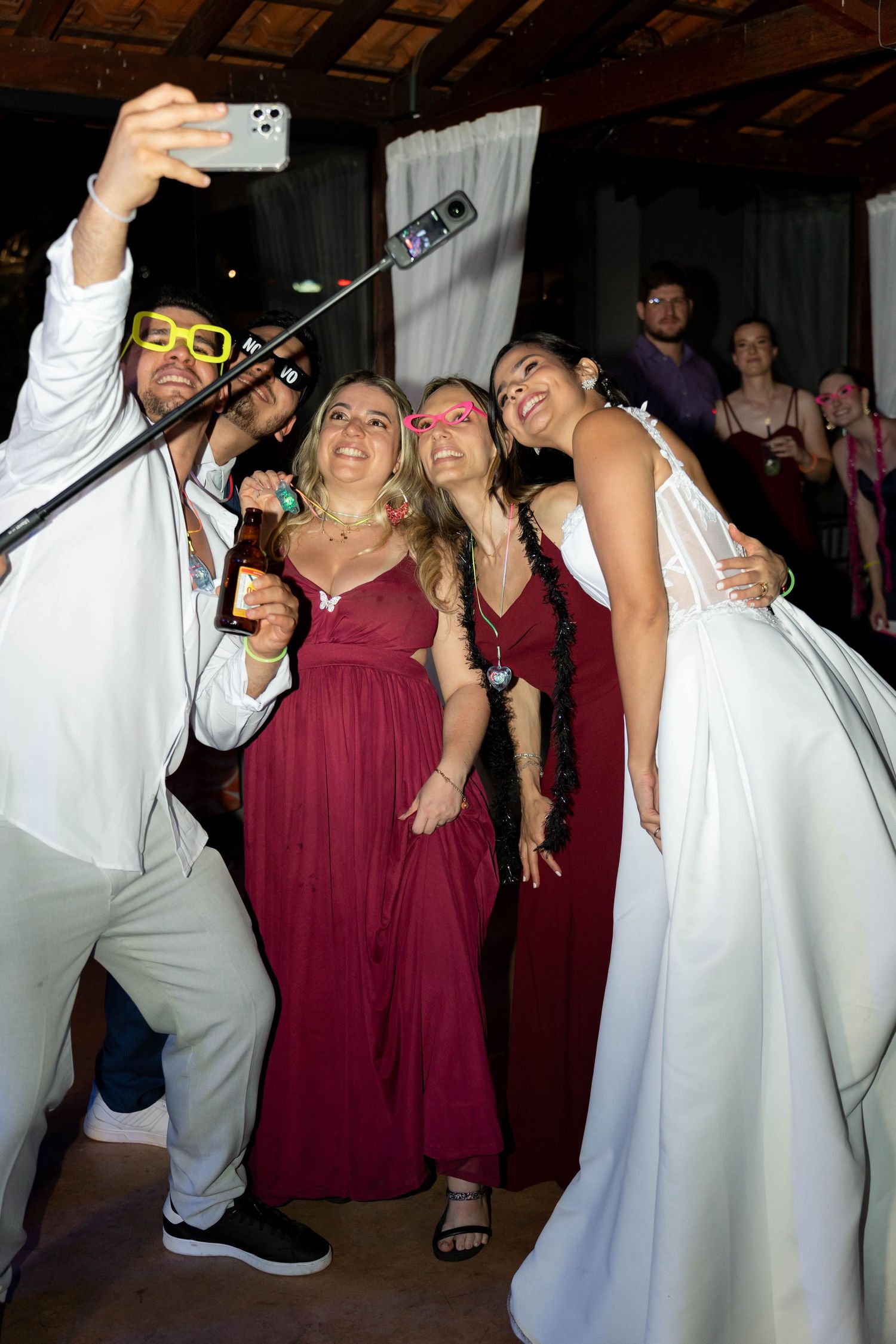 A bride in white and bridesmaids in burgundy dresses take a selfie with a selfie stick at a wedding reception.