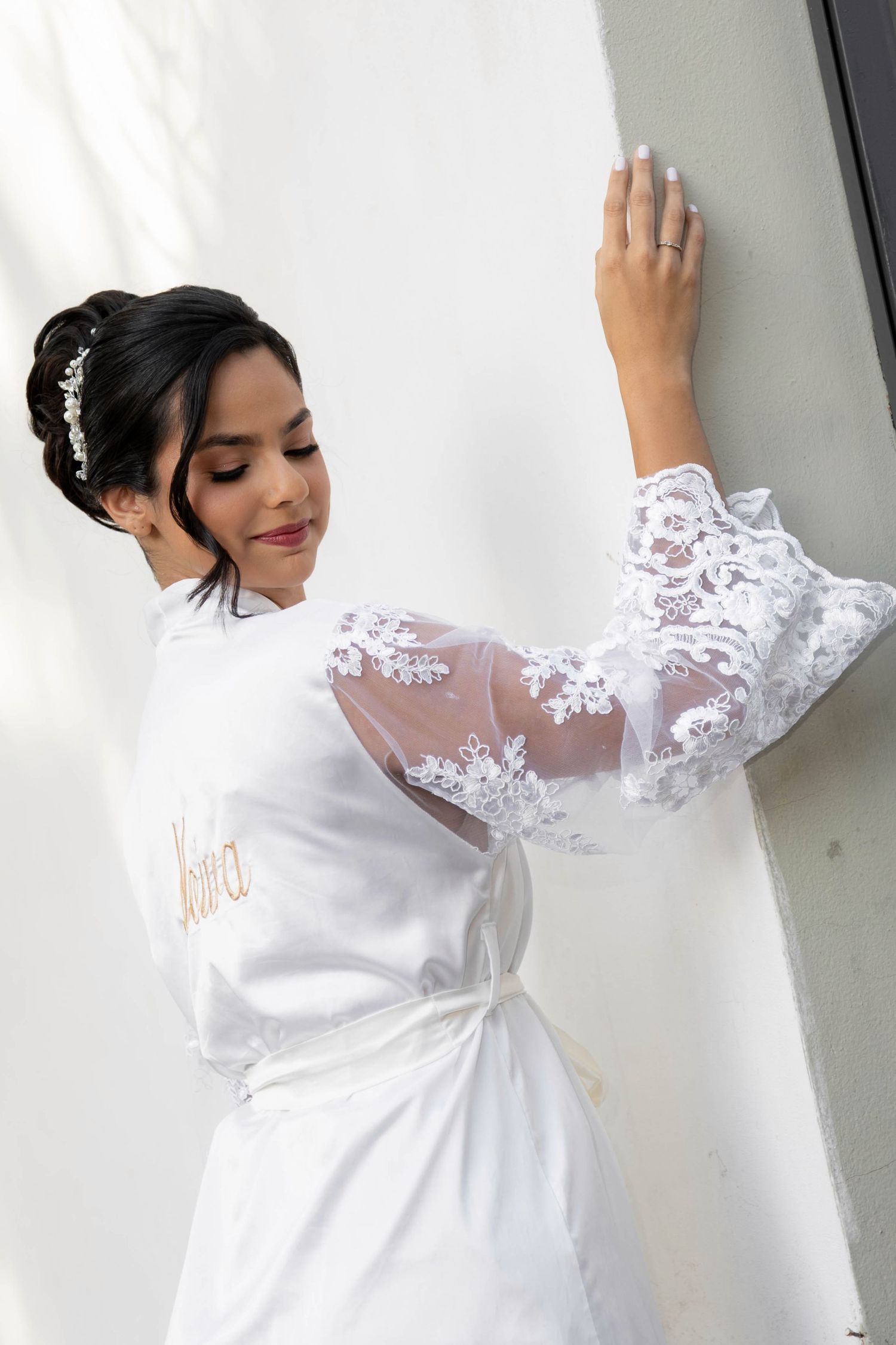 Bride in white lace robe with sheer sleeves, hair up with floral accessory, posing gracefully against a white wall.