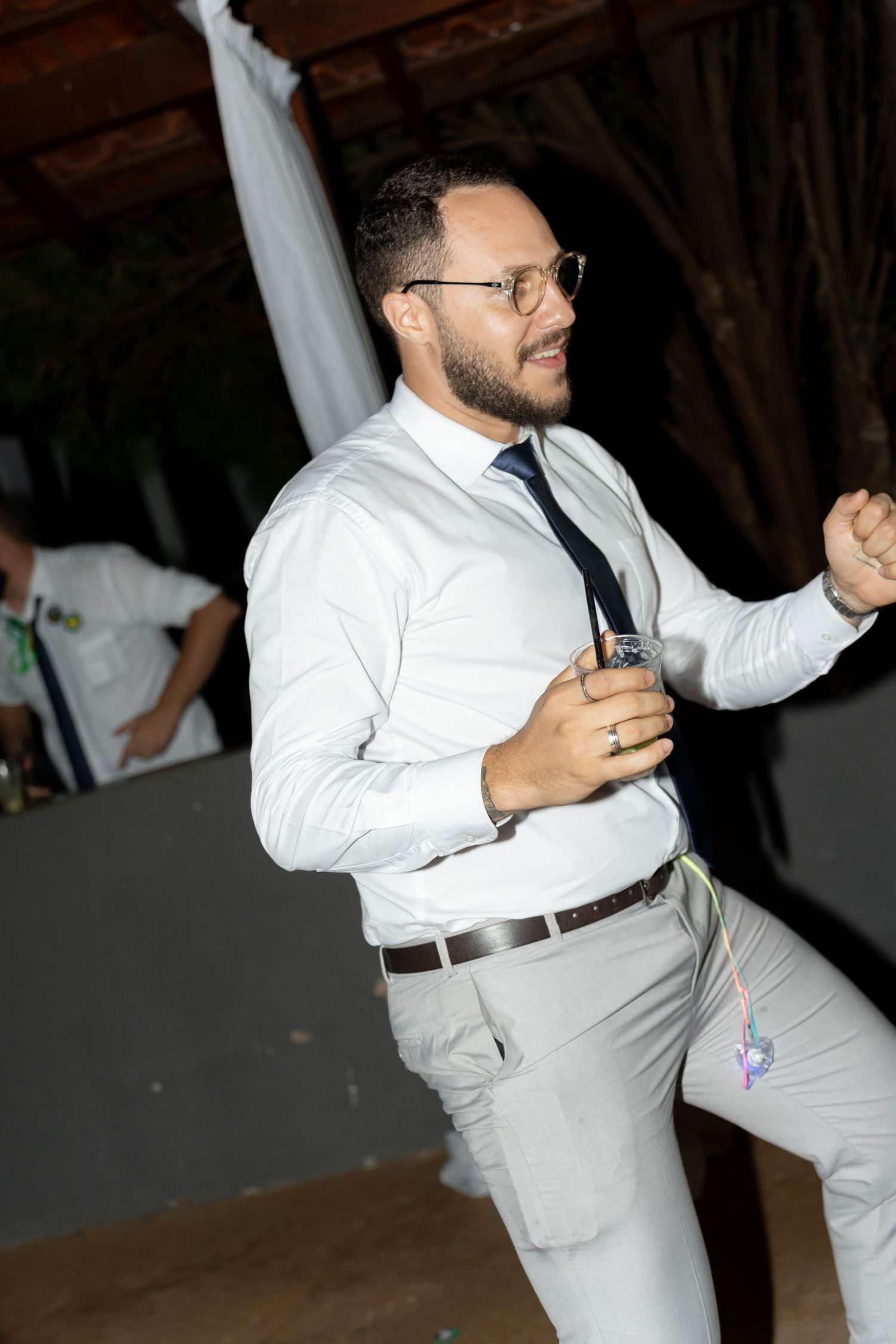 Man in white shirt and tie dancing at a formal event, holding a drink and smiling.