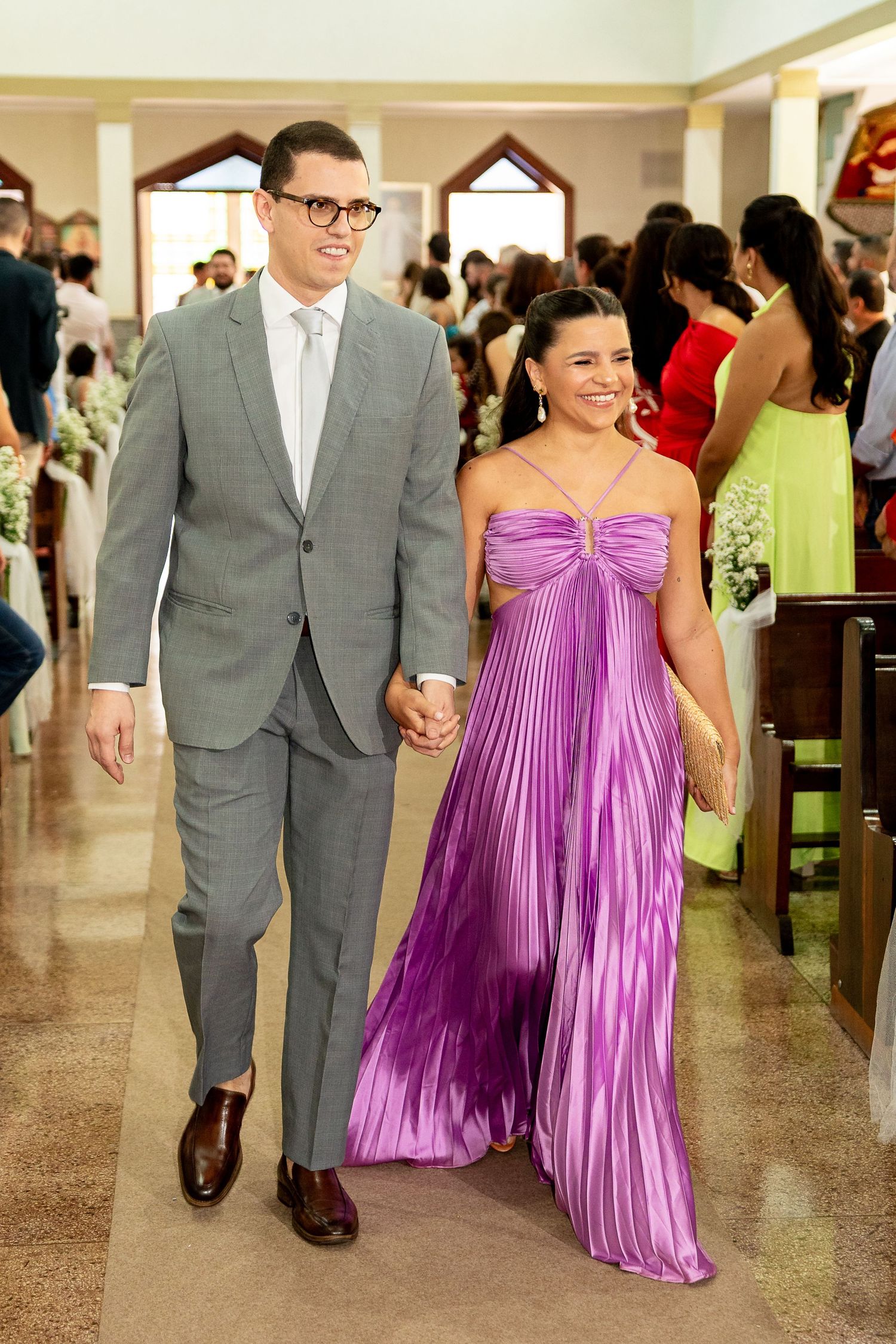 Couple walking down church aisle, man in gray suit, woman in purple pleated gown, wedding ceremony.