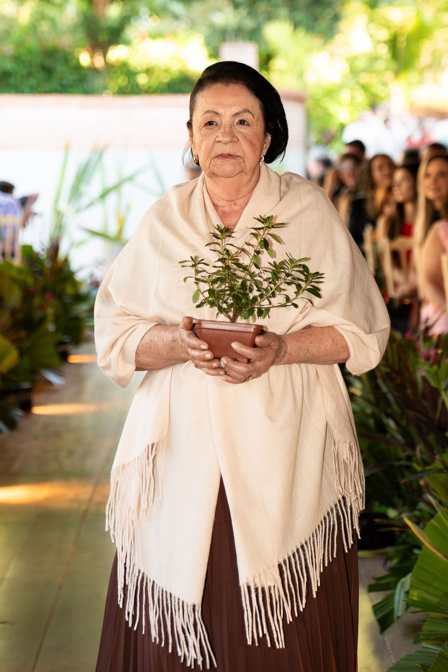 Elderly woman in white shawl holding a potted plant walks down an aisle lined with greenery.