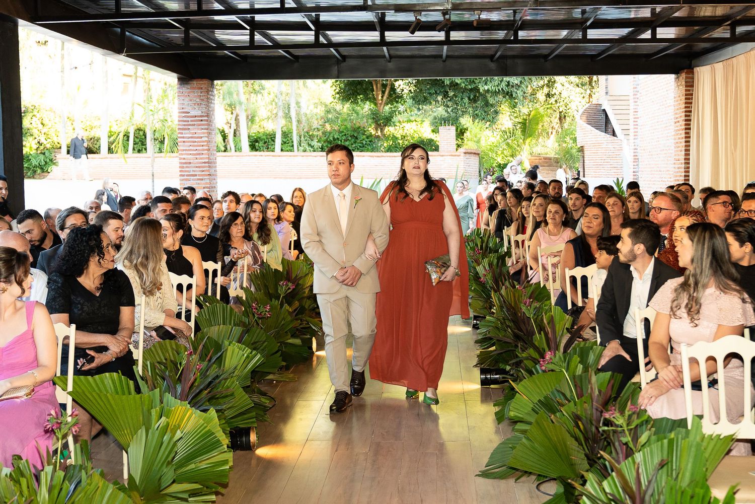 Couple walks down wedding aisle surrounded by guests and tropical greenery at an elegant outdoor ceremony.