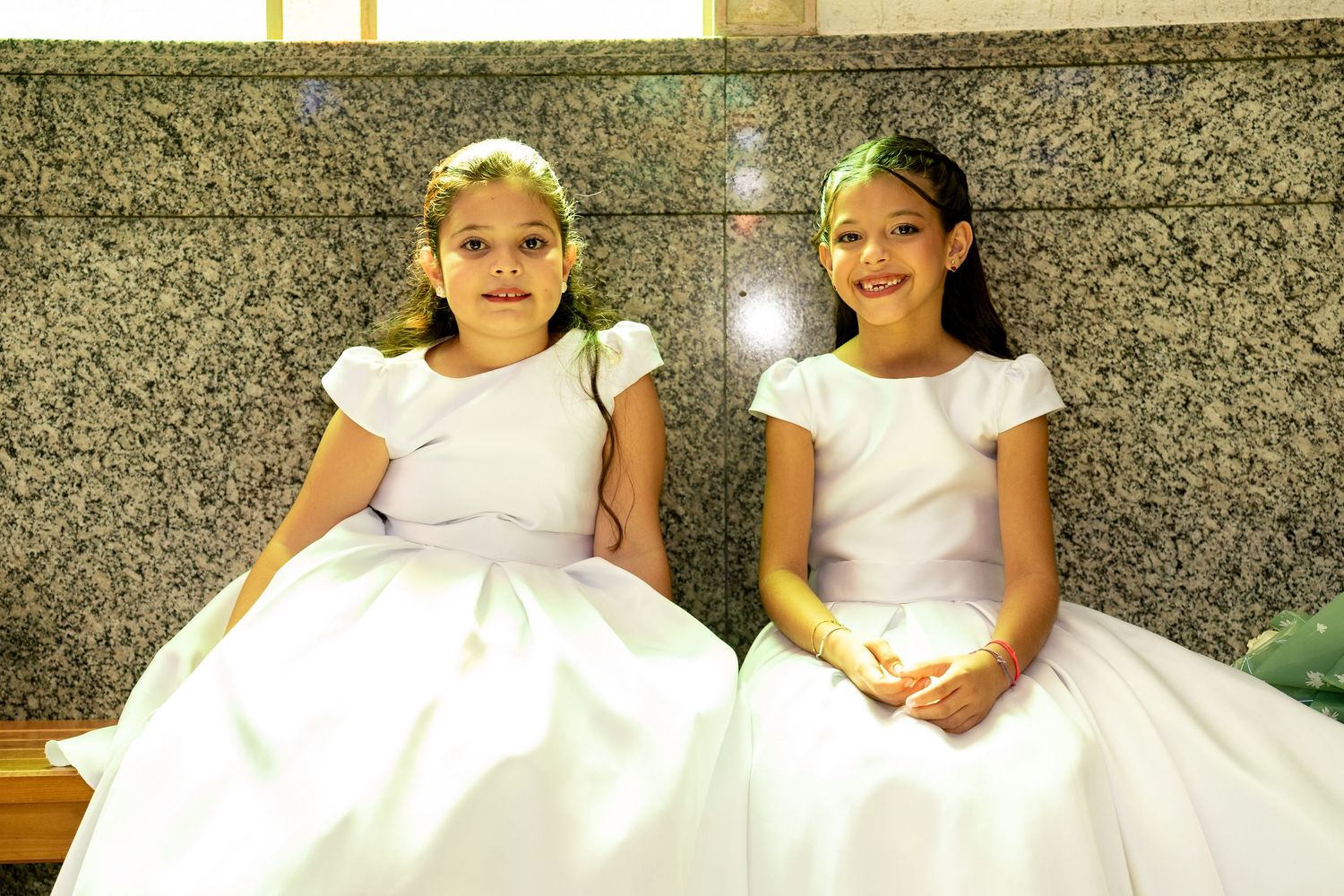 Two young girls in matching white dresses sitting together, smiling at a formal religious ceremony.